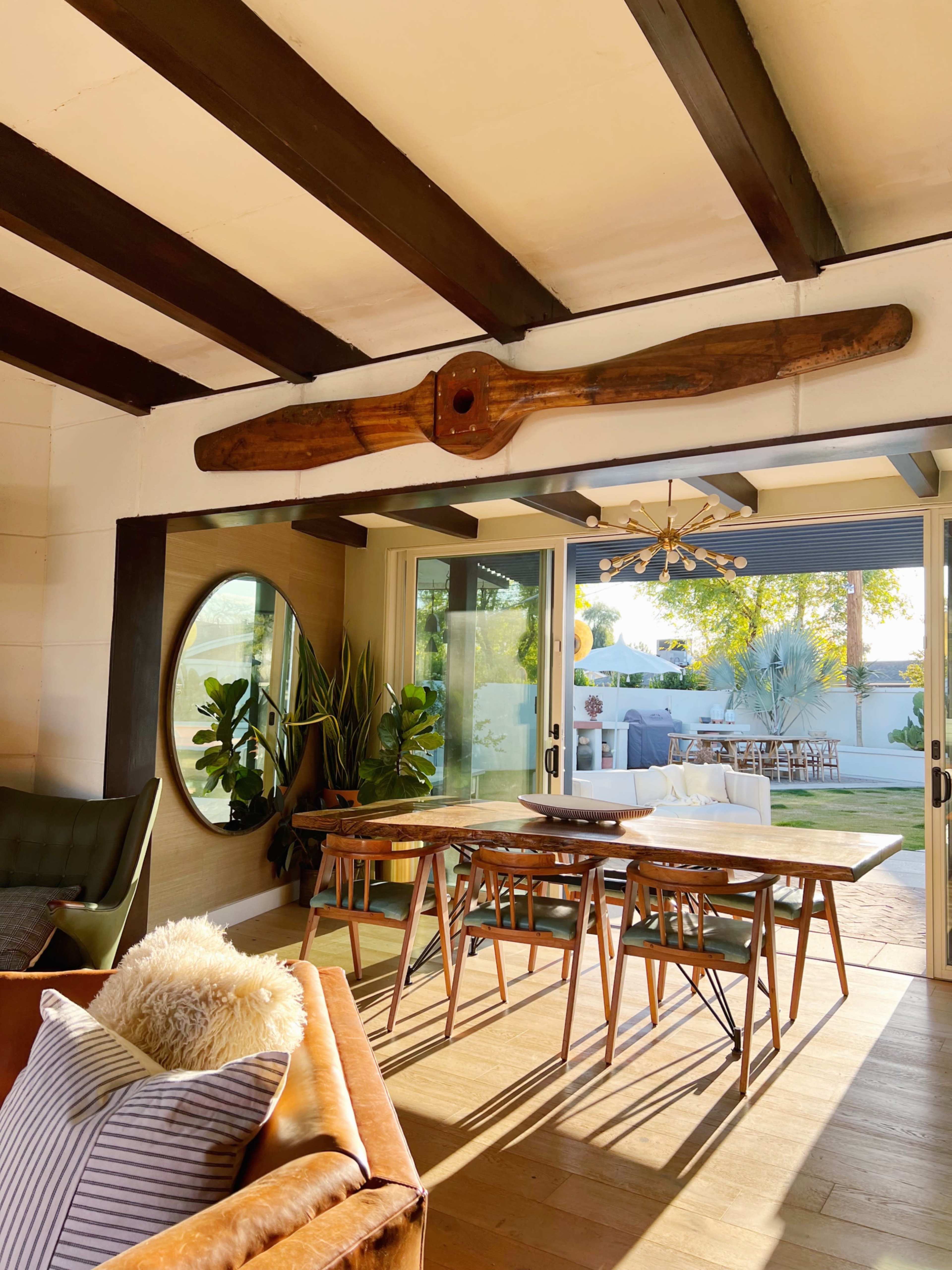 The image shows a modern interior space featuring a wooden beam, a dining table with chairs, and large glass doors that open to an outdoor area.