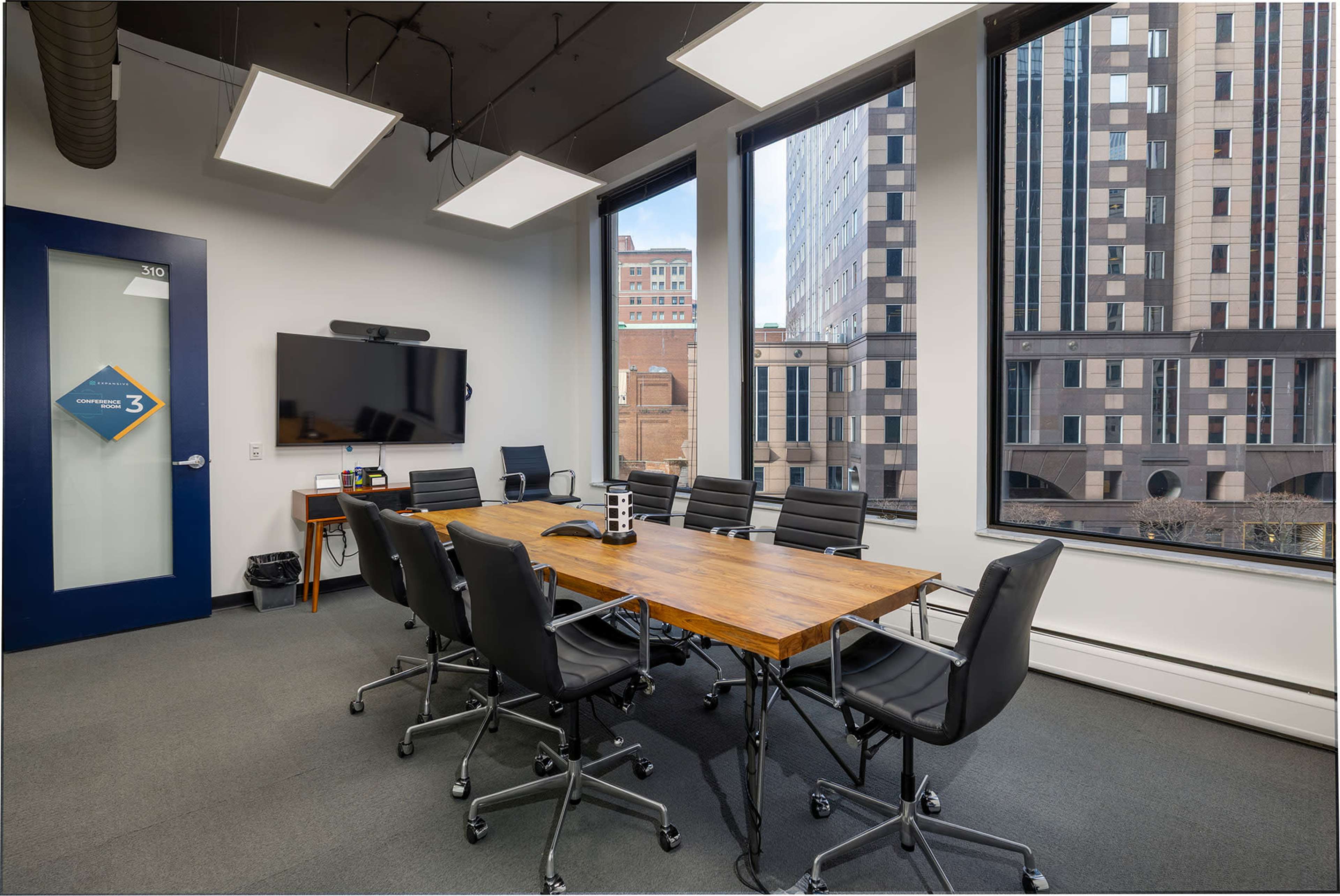 The image shows a modern conference room featuring a large wooden table surrounded by black chairs, with large windows revealing a cityscape outside.