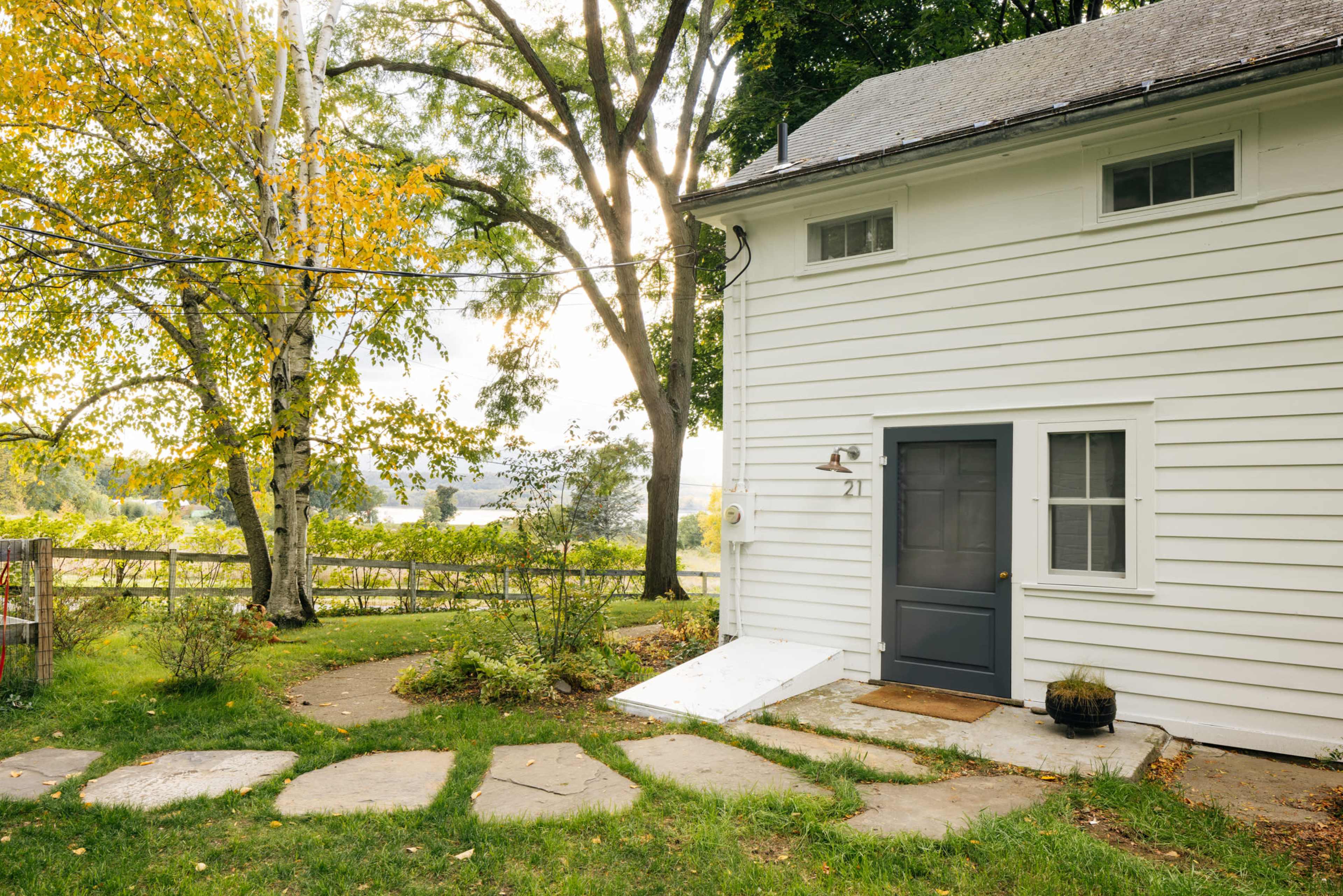A gray house with white siding has a main entrance door and a stone pathway leading to it, surrounded by trees and a grassy area.