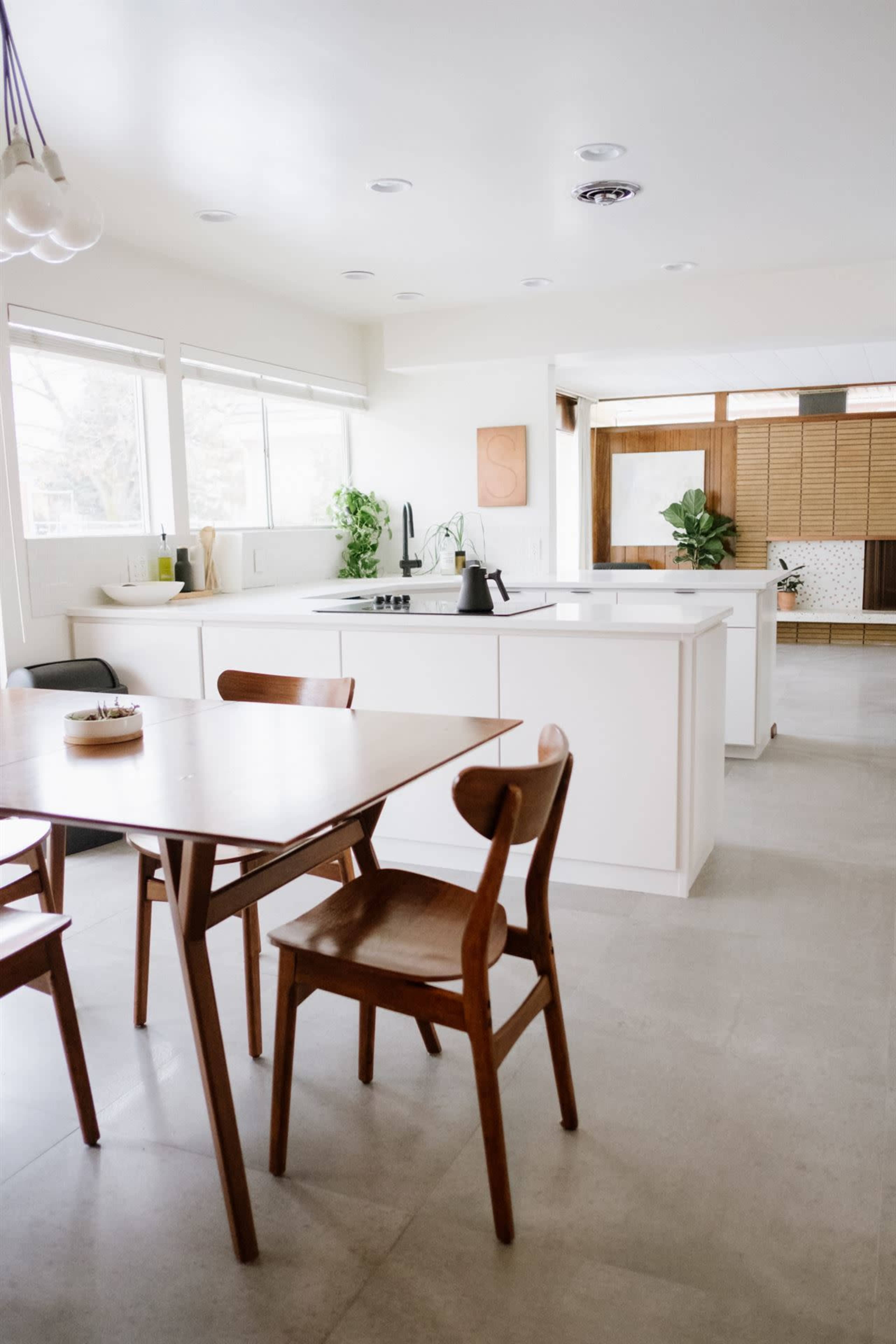 A modern kitchen features a wooden dining table with chairs, a sink, and plants by the windows, with light-colored cabinets and a spacious layout.