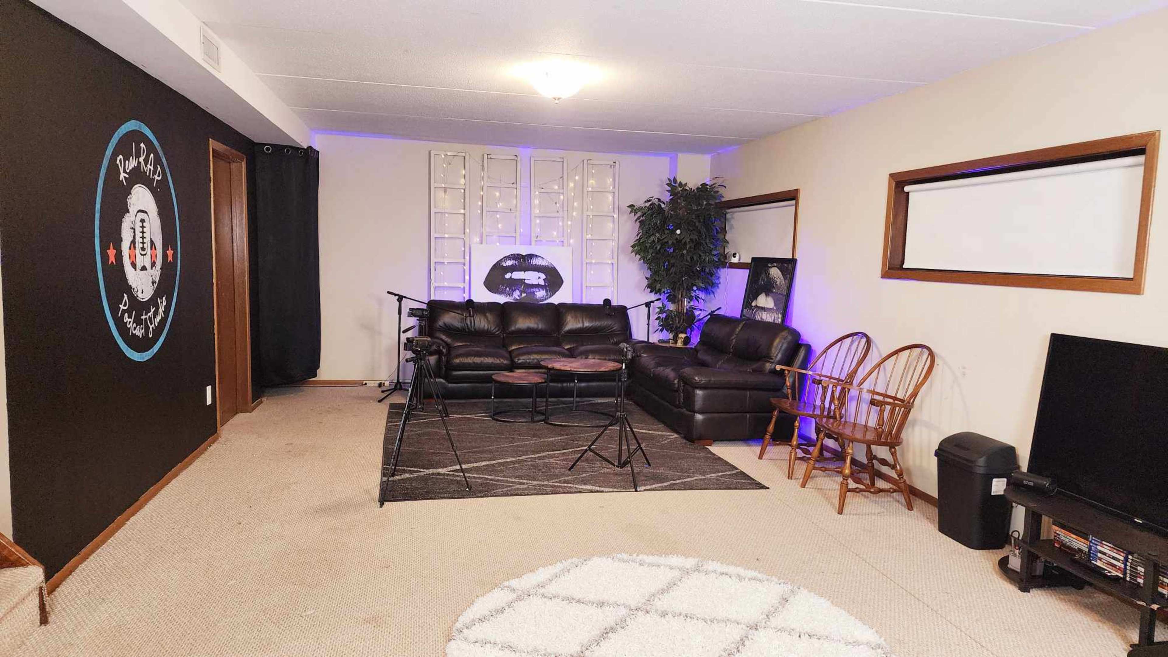 The image shows a living room with dark walls, a sectional couch, two wooden chairs, a television, and a musical setup with microphones and stands.