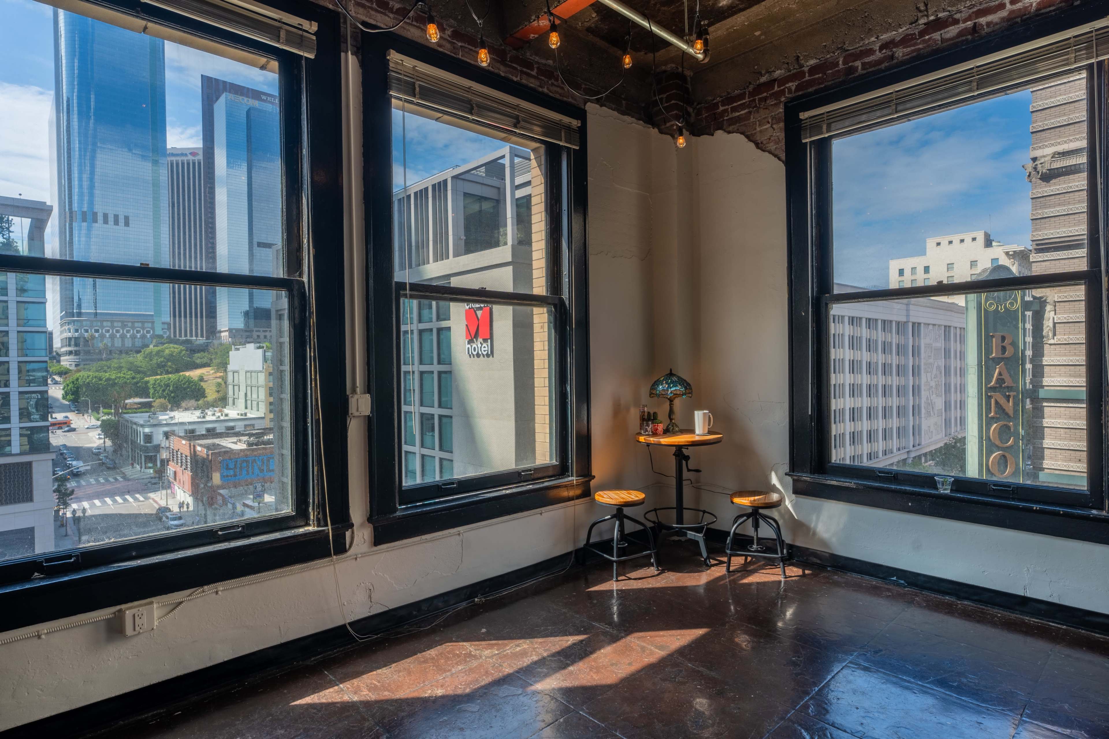 A corner of a room with two large windows overlooking a cityscape, featuring two stools and a small table in the foreground.
