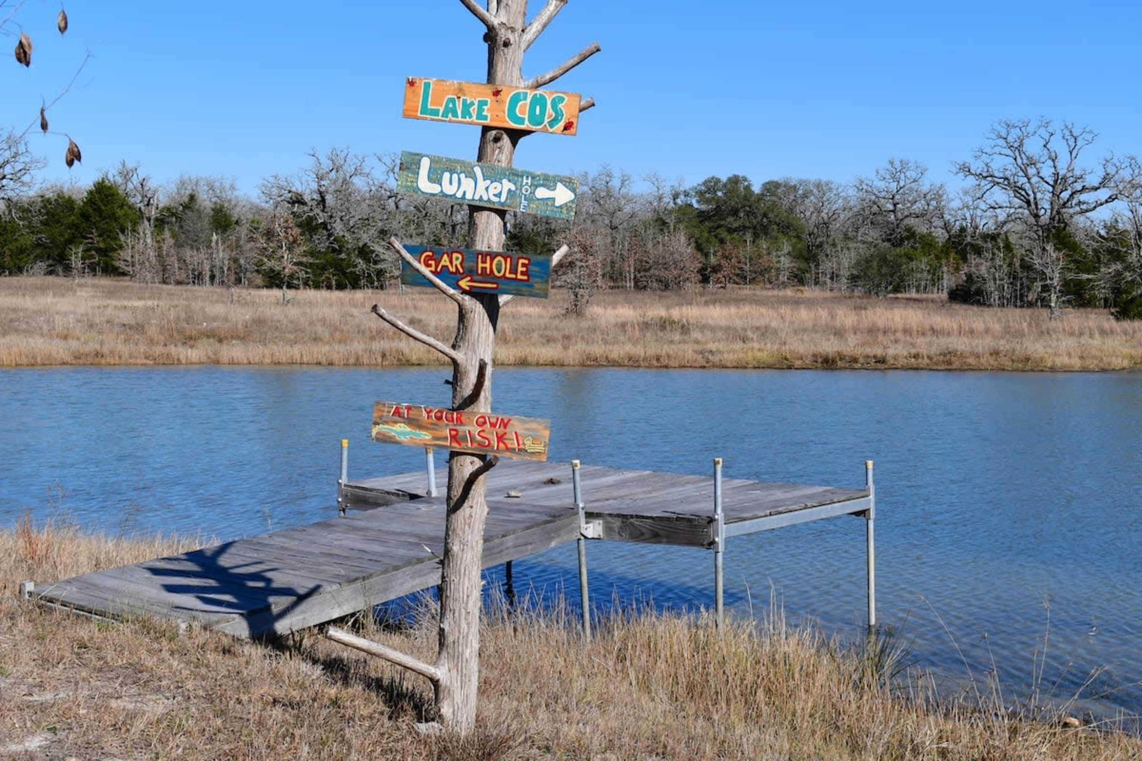 A wooden signpost with multiple directional signs stands near a dock by a calm lake, surrounded by dry grass and sparse trees.