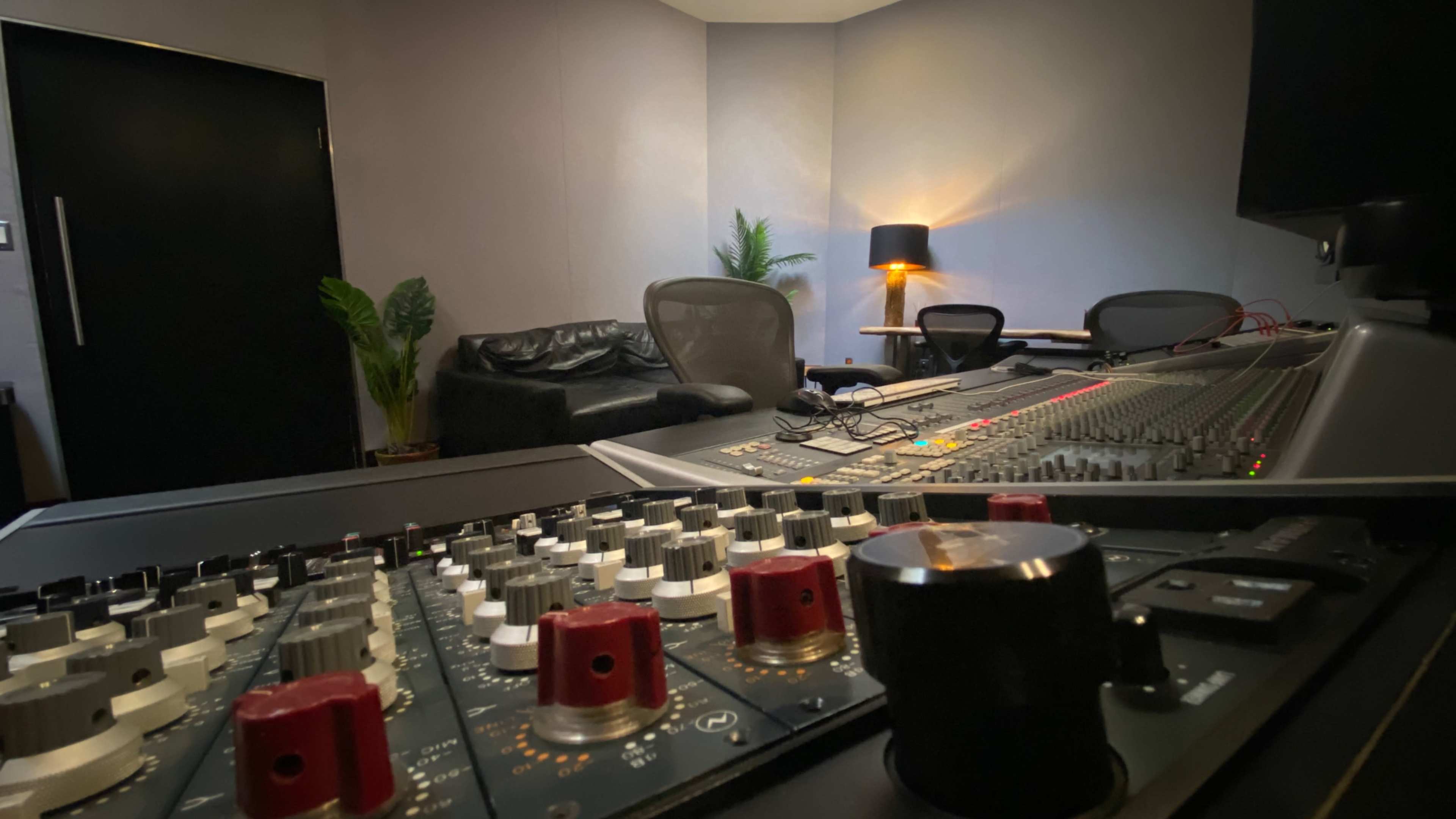 A recording studio is shown with a large mixing console in the foreground and a cozy seating area in the background.