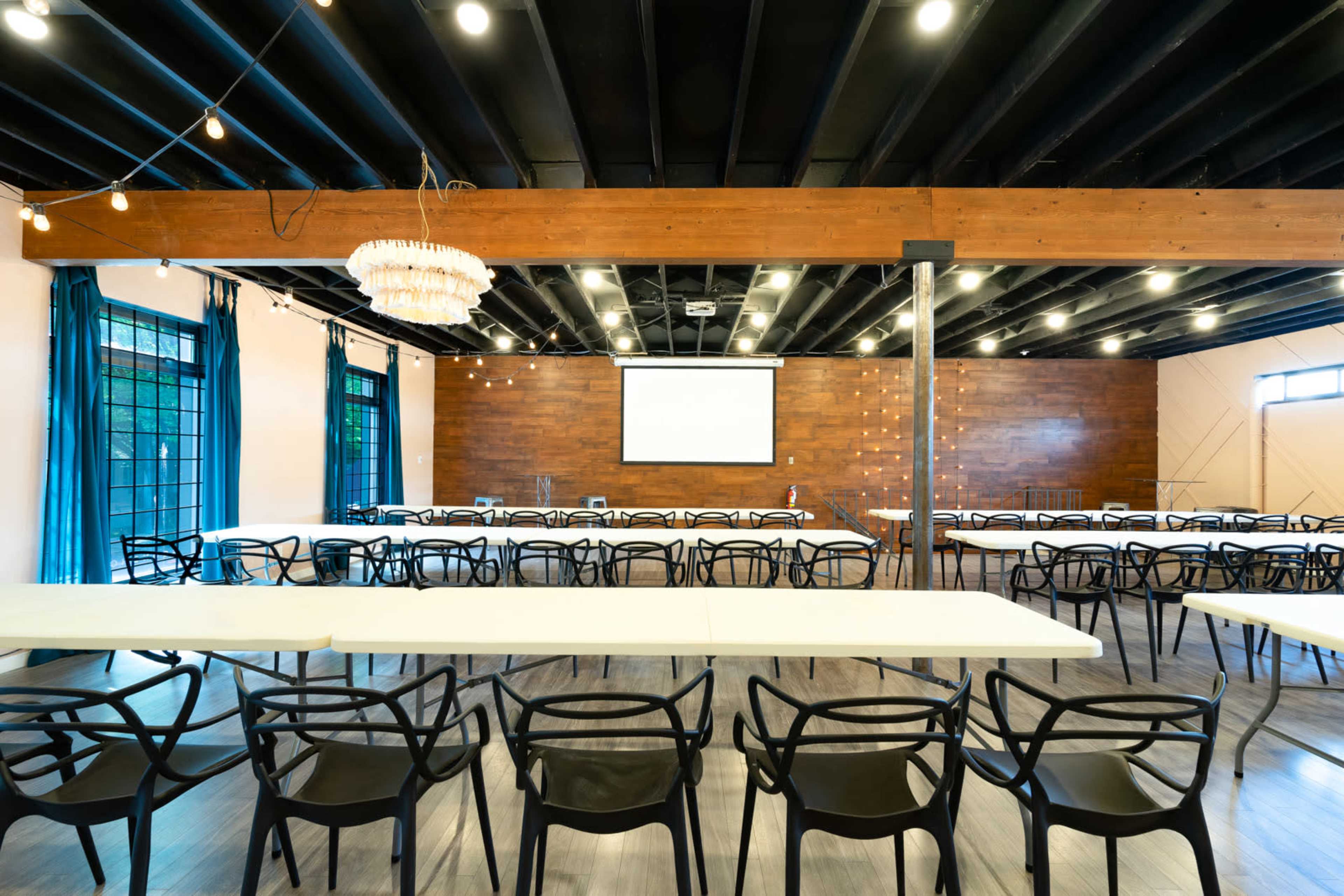 A spacious event room set up with rows of tables and chairs, featuring wooden accents and a projector screen at one end.