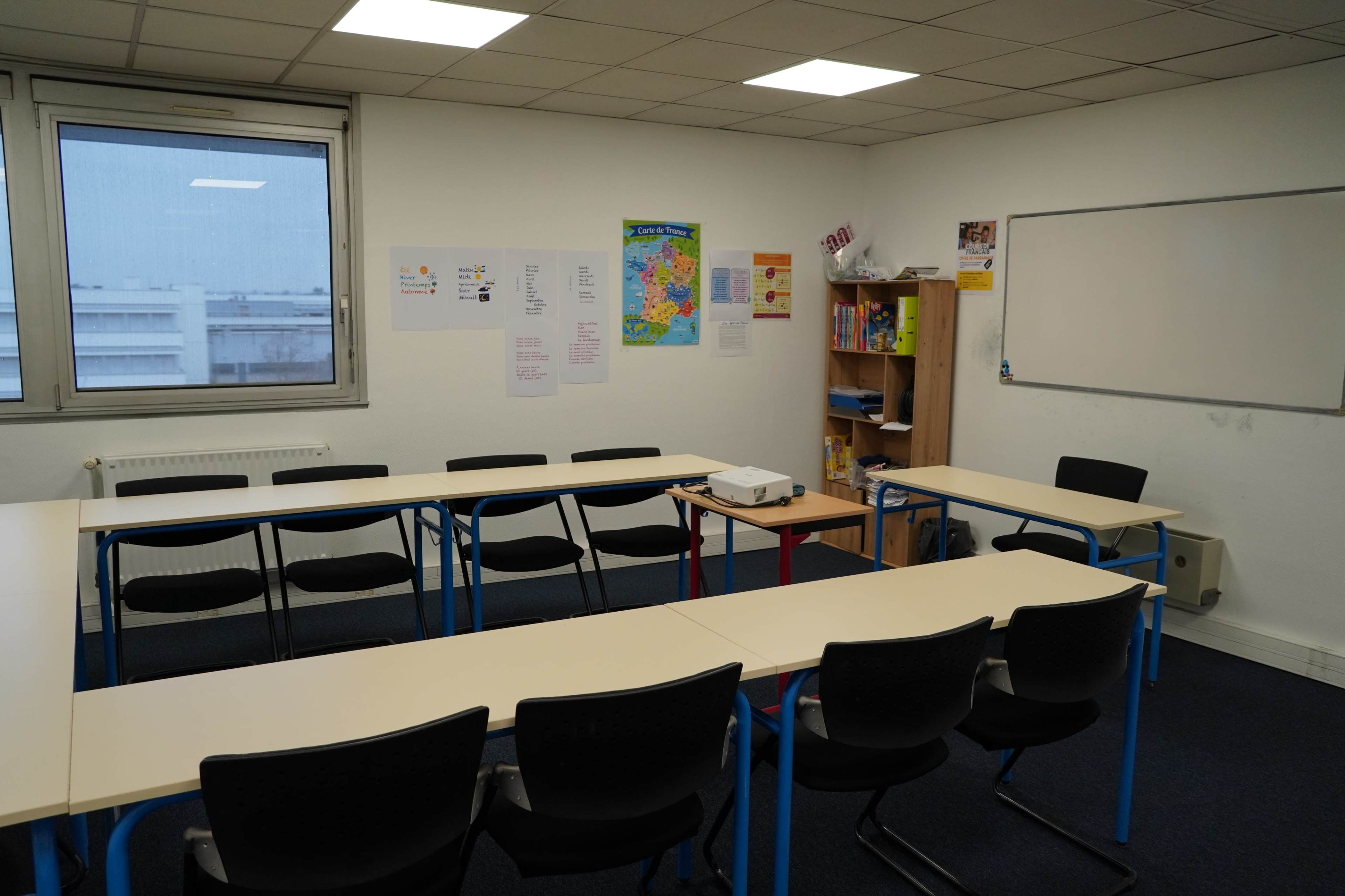 The image shows a classroom equipped with tables arranged in a U-shape, a projector on a low table, a bookshelf filled with materials, and educational posters on the walls.