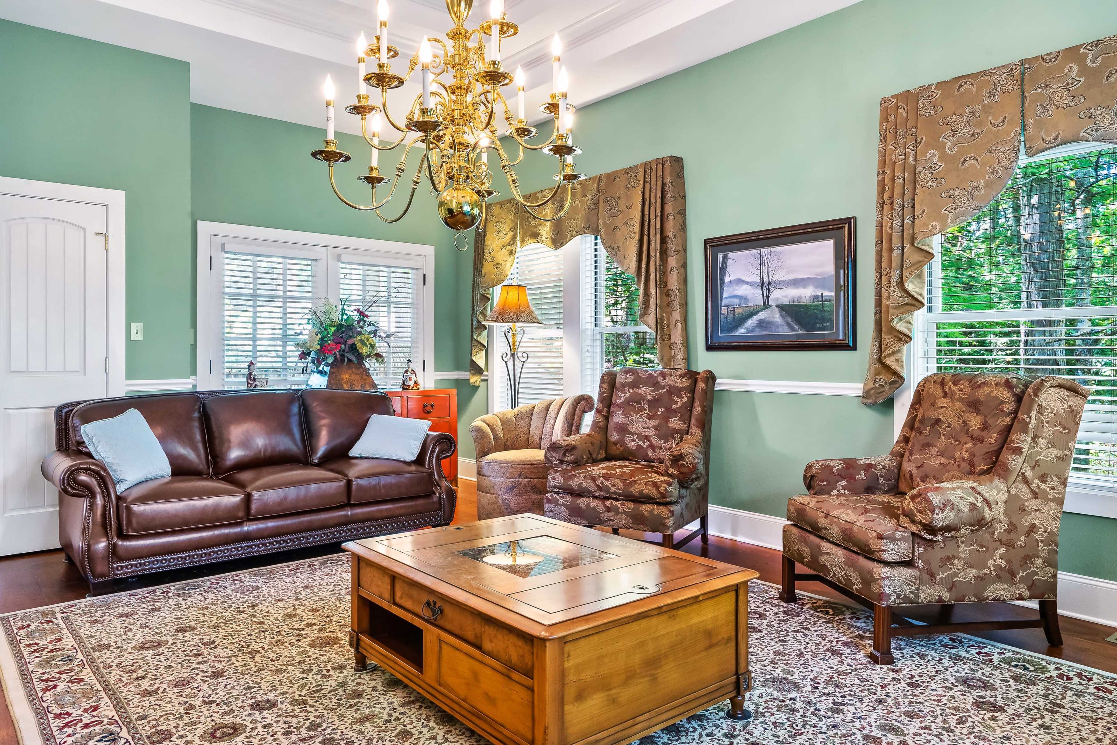 A living room with a brown leather sofa, two upholstered armchairs, a wooden coffee table, and a chandelier, against a green wall and large windows with drapes.