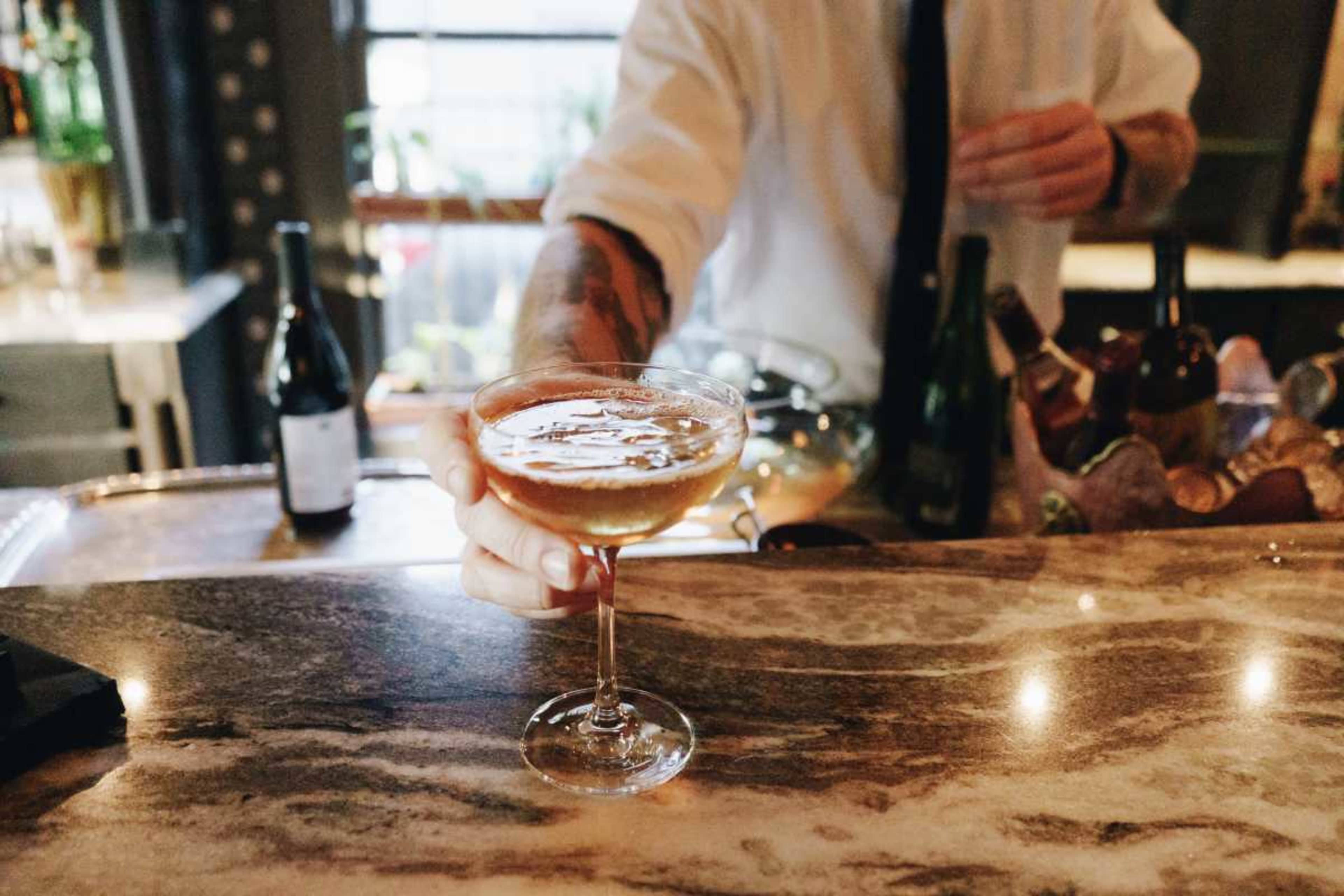A bartender is serving a cocktail in a coupe glass over a polished bar countertop.
