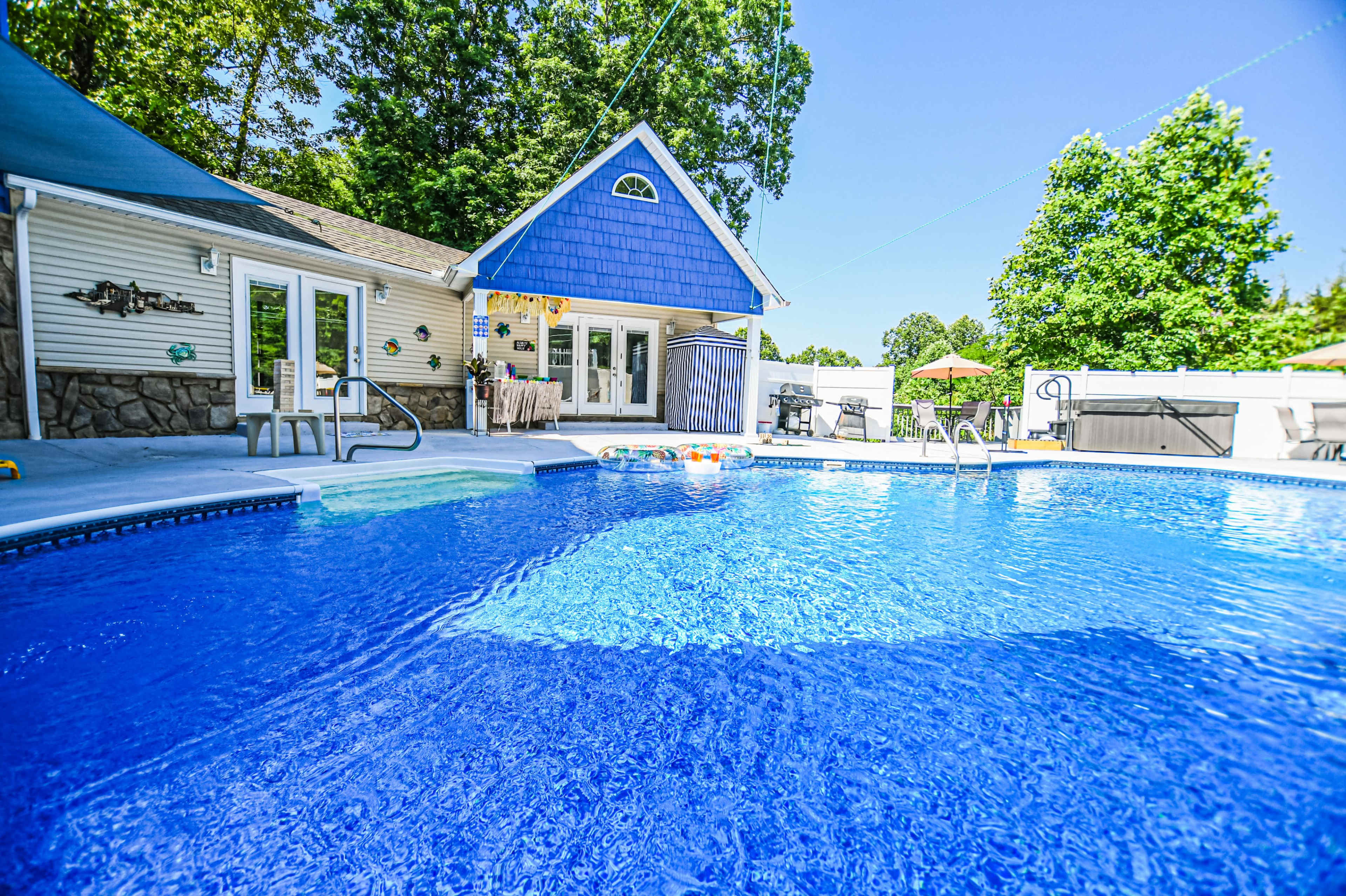 A backyard swimming pool surrounded by a house, lounge chairs, and green trees under a clear blue sky.