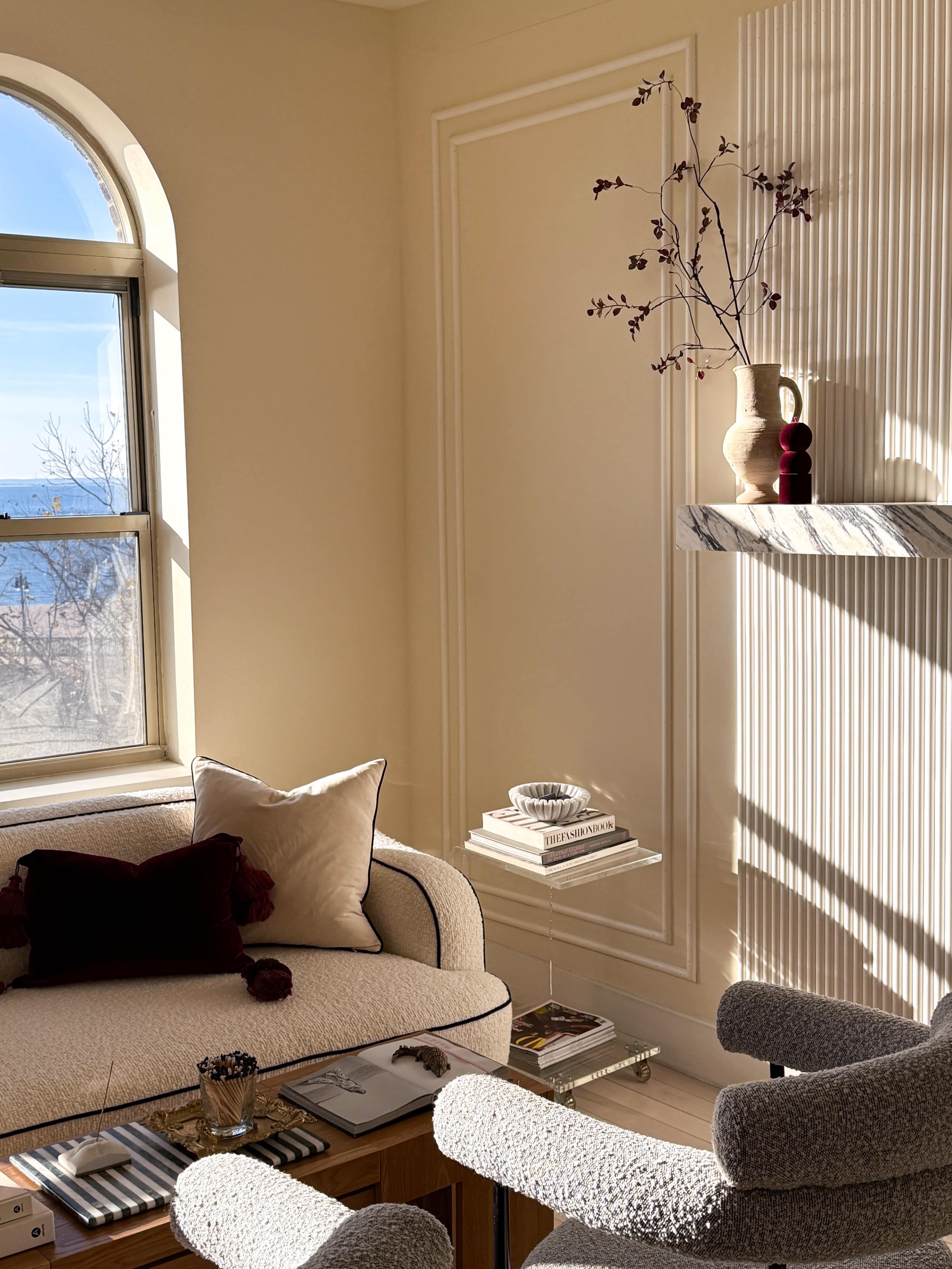 A cozy living room features a comfortable sofa with decorative pillows, a clear glass side table, and a marble shelf displaying a vase with dried flowers.