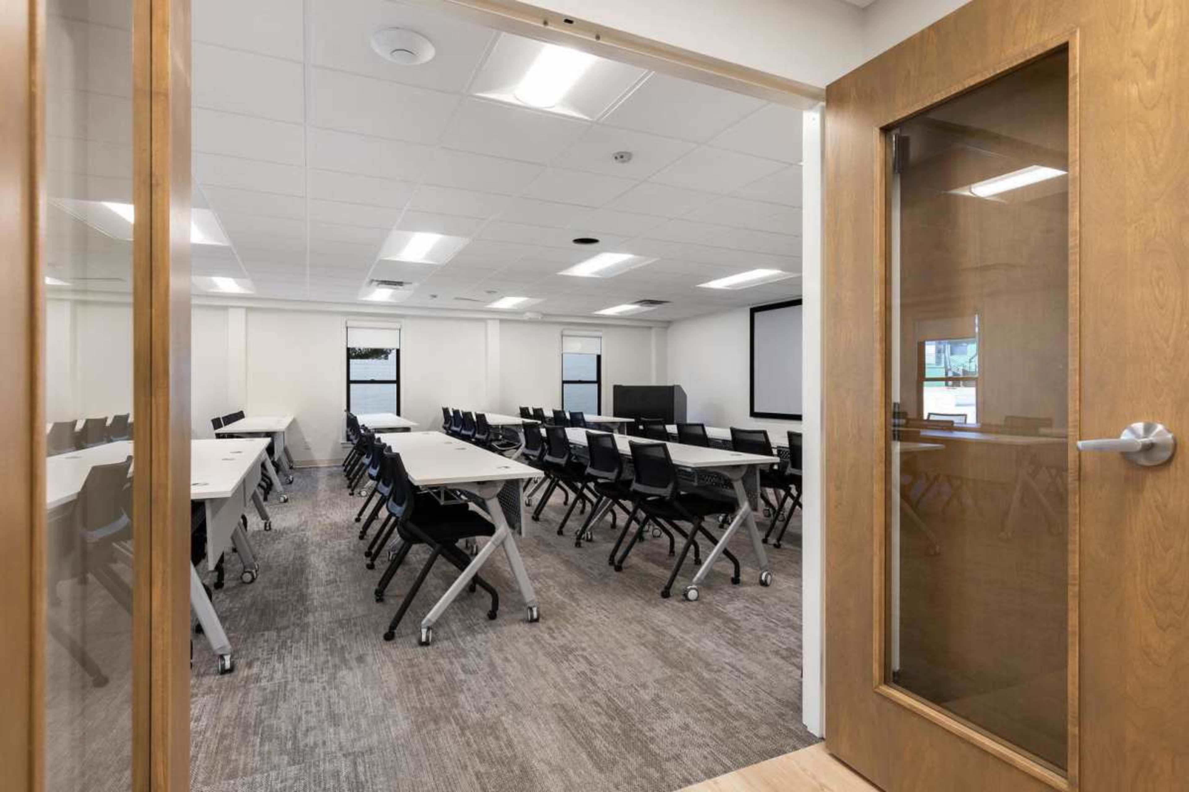 The image shows a modern classroom with rows of tables and chairs arranged for a meeting or educational session, viewed from an open doorway.