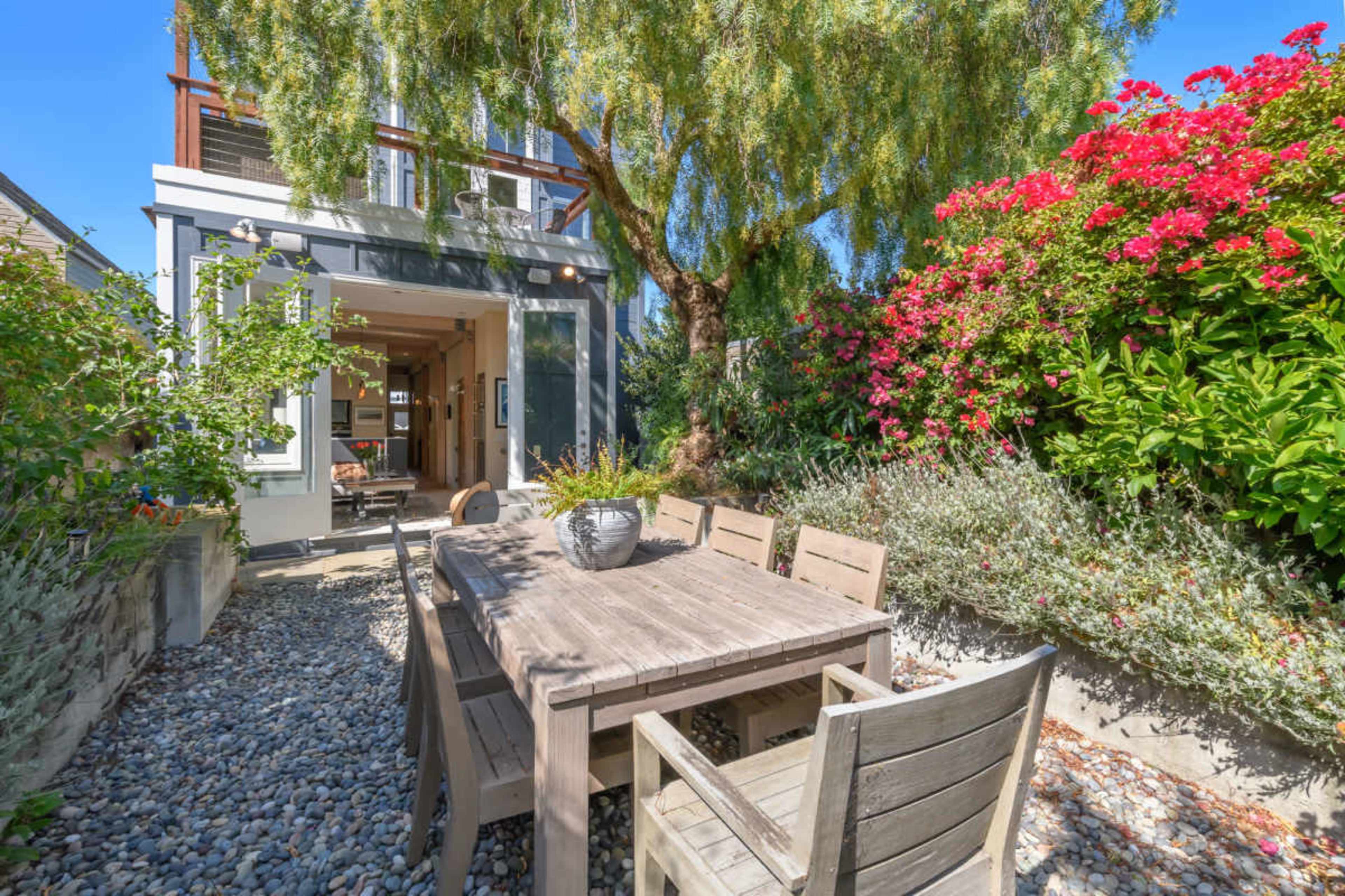 A wooden dining table with chairs is set in a stone garden, surrounded by flowering shrubs and a tree, with a modern house visible in the background.