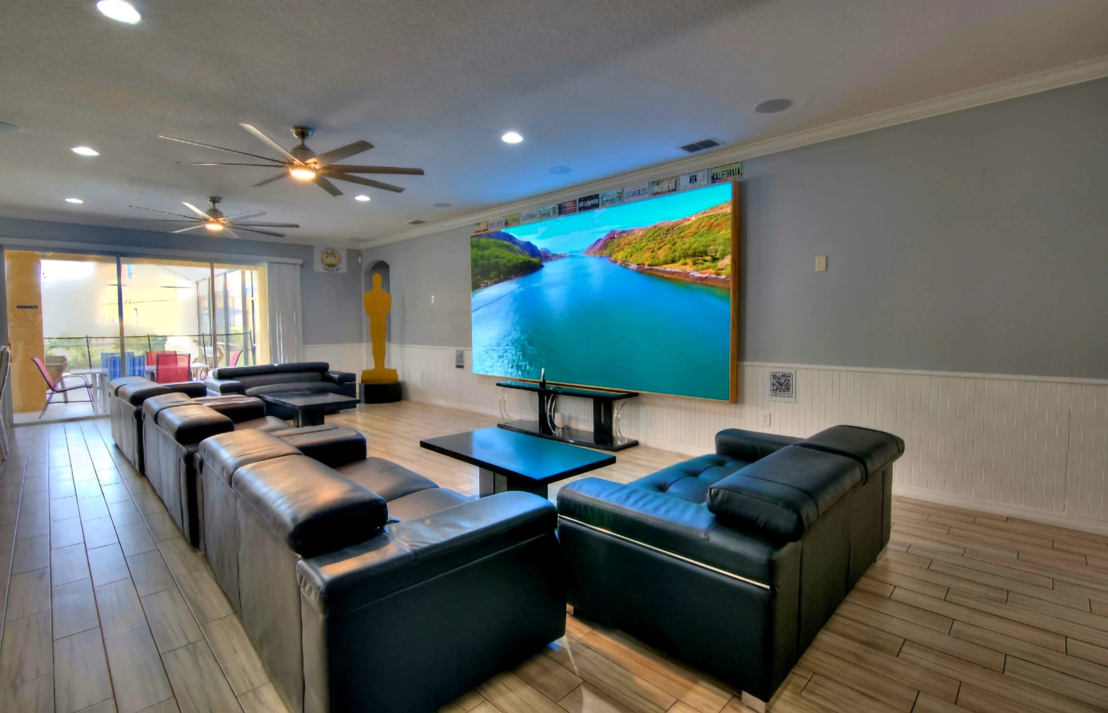 A spacious living room features black leather sofas facing a large screen displaying a scenic view of a river and mountains.