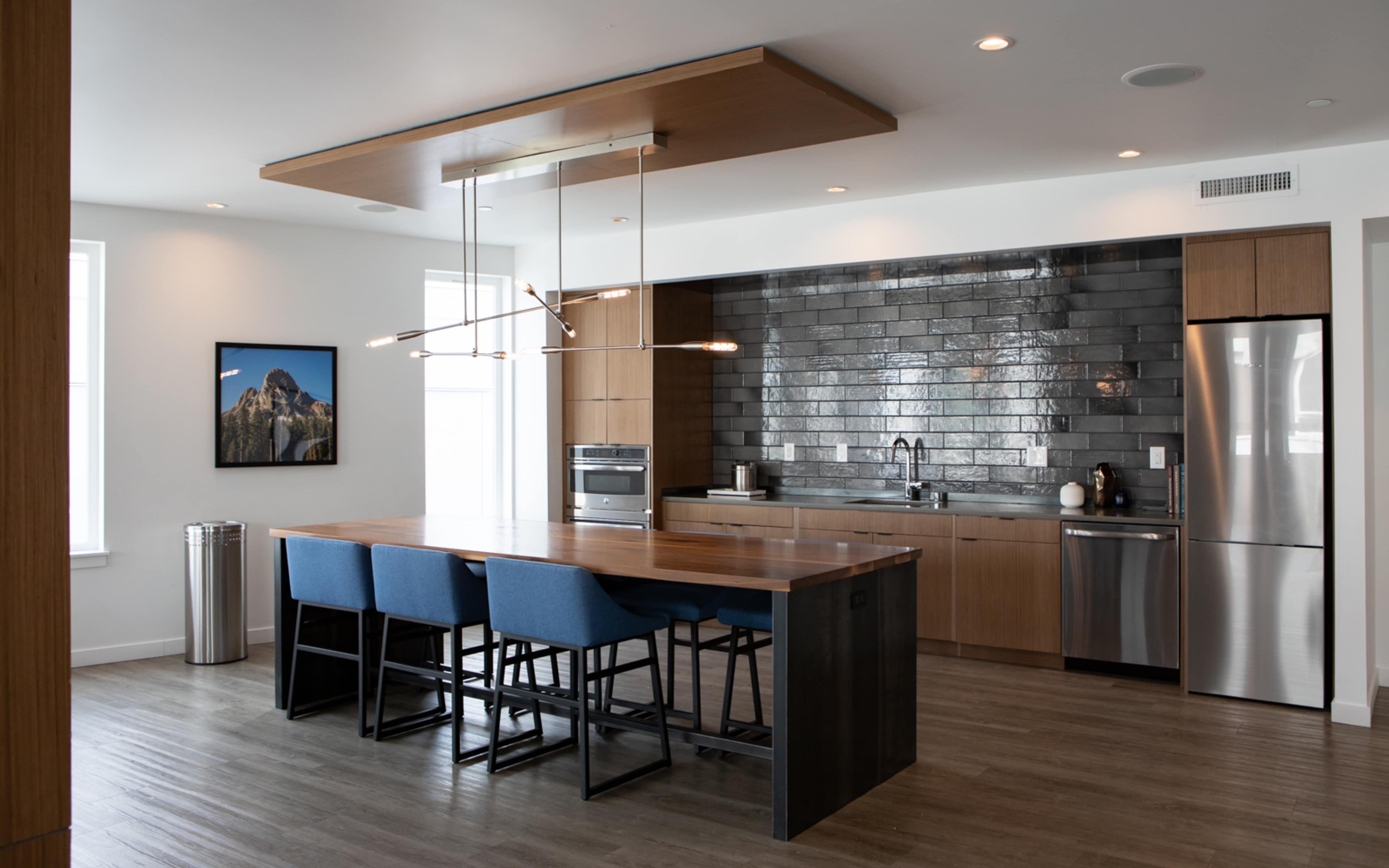 A modern kitchen with a wooden island, blue chairs, stainless steel appliances, and a gray tiled backsplash.