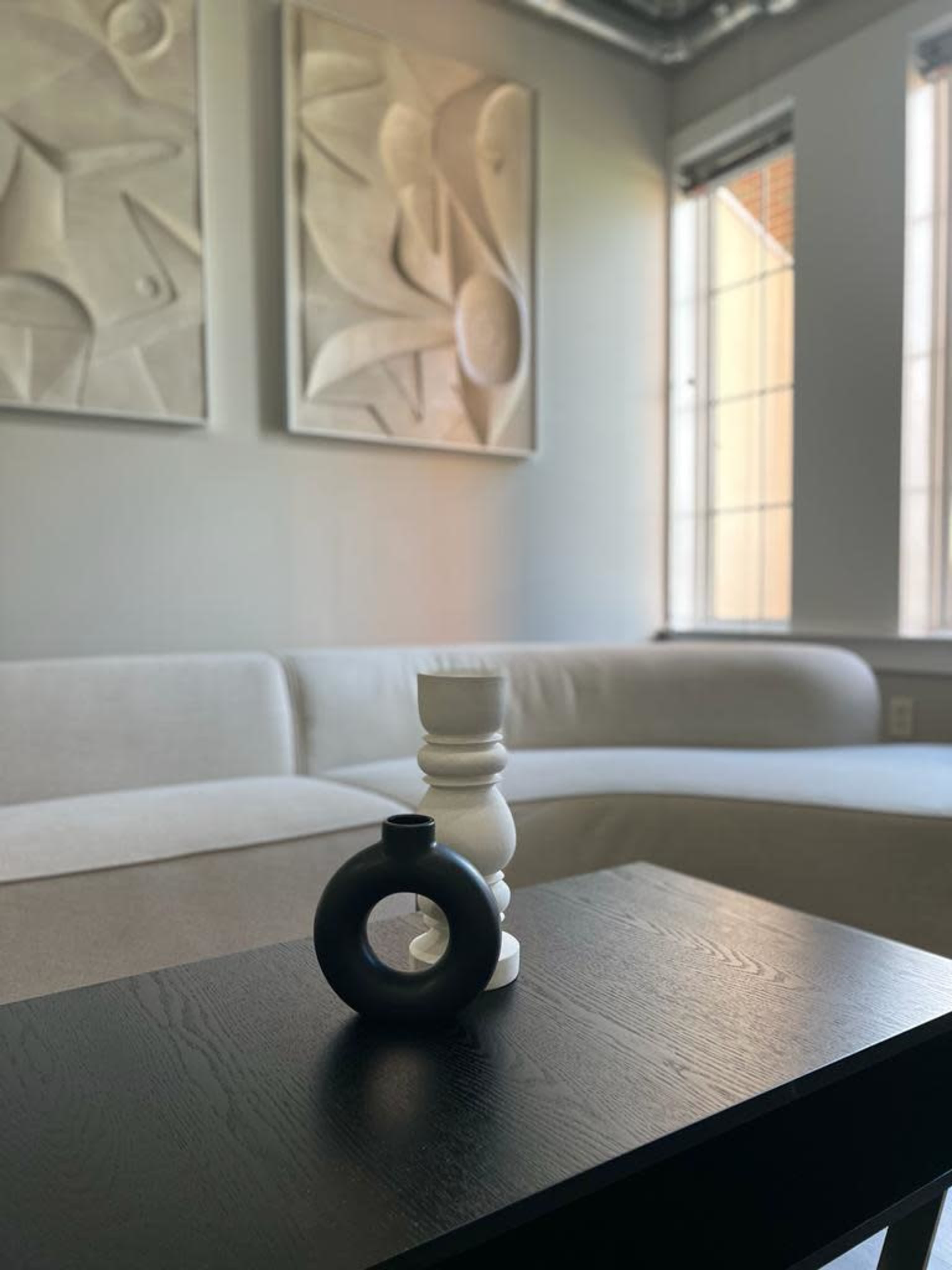 A white and black decorative vase and a candle holder are placed on a dark wooden table in a minimalist living room with abstract wall art and a sectional sofa.