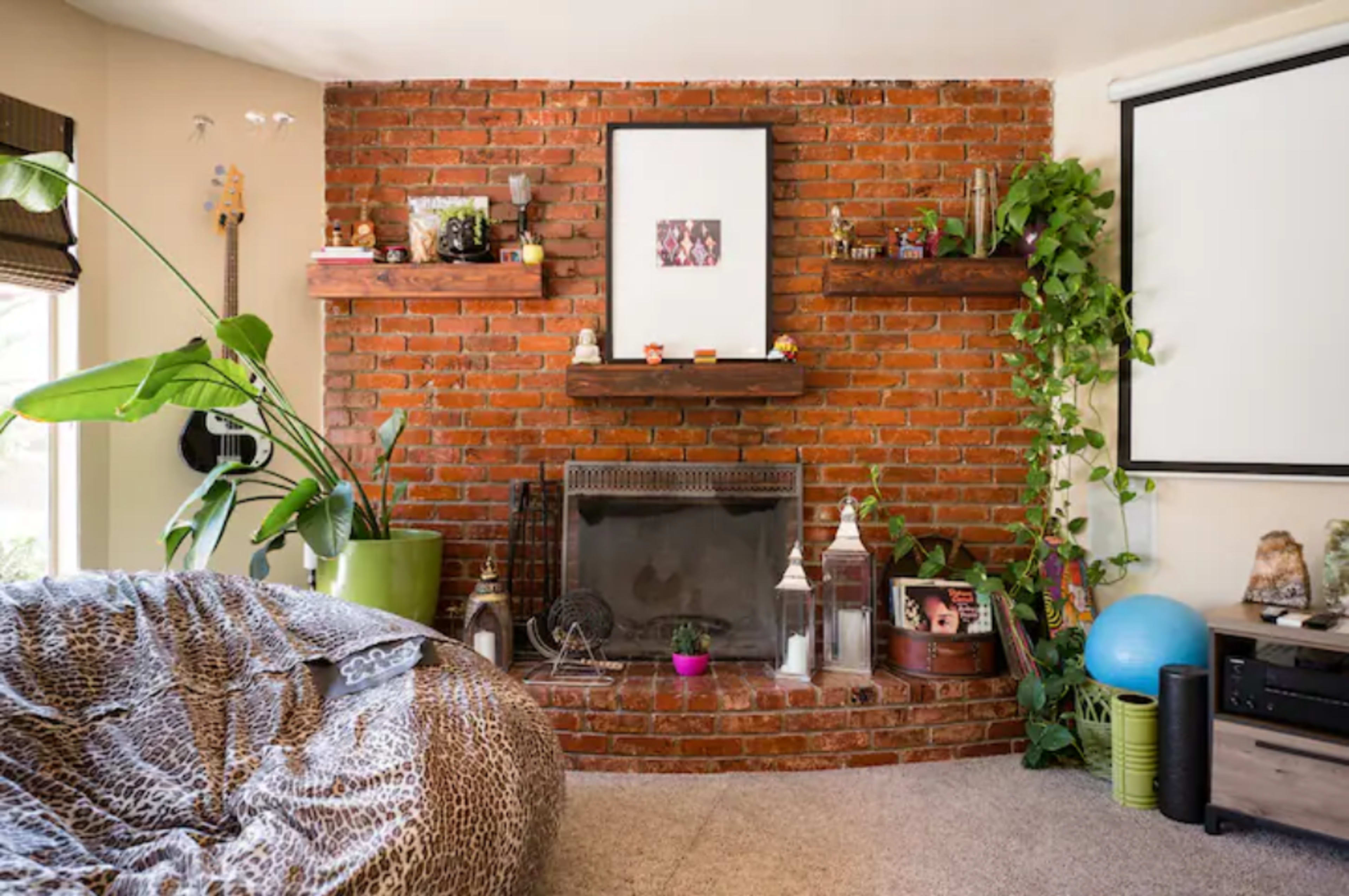 A cozy living room with a brick wall, a fireplace, shelves adorned with decorations, a large bean bag chair, and various plants.