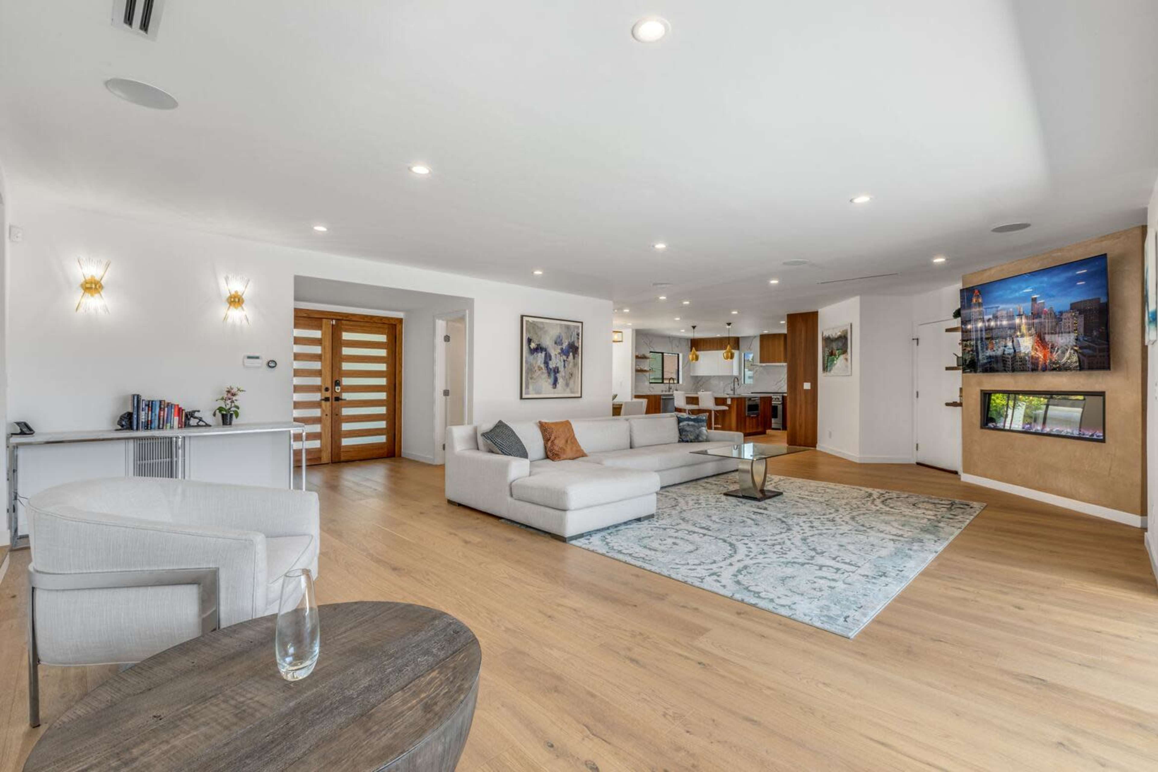 A spacious living room features a light-colored sectional sofa, a round coffee table, and a large wall-mounted television, with an open layout connecting to a kitchen area.