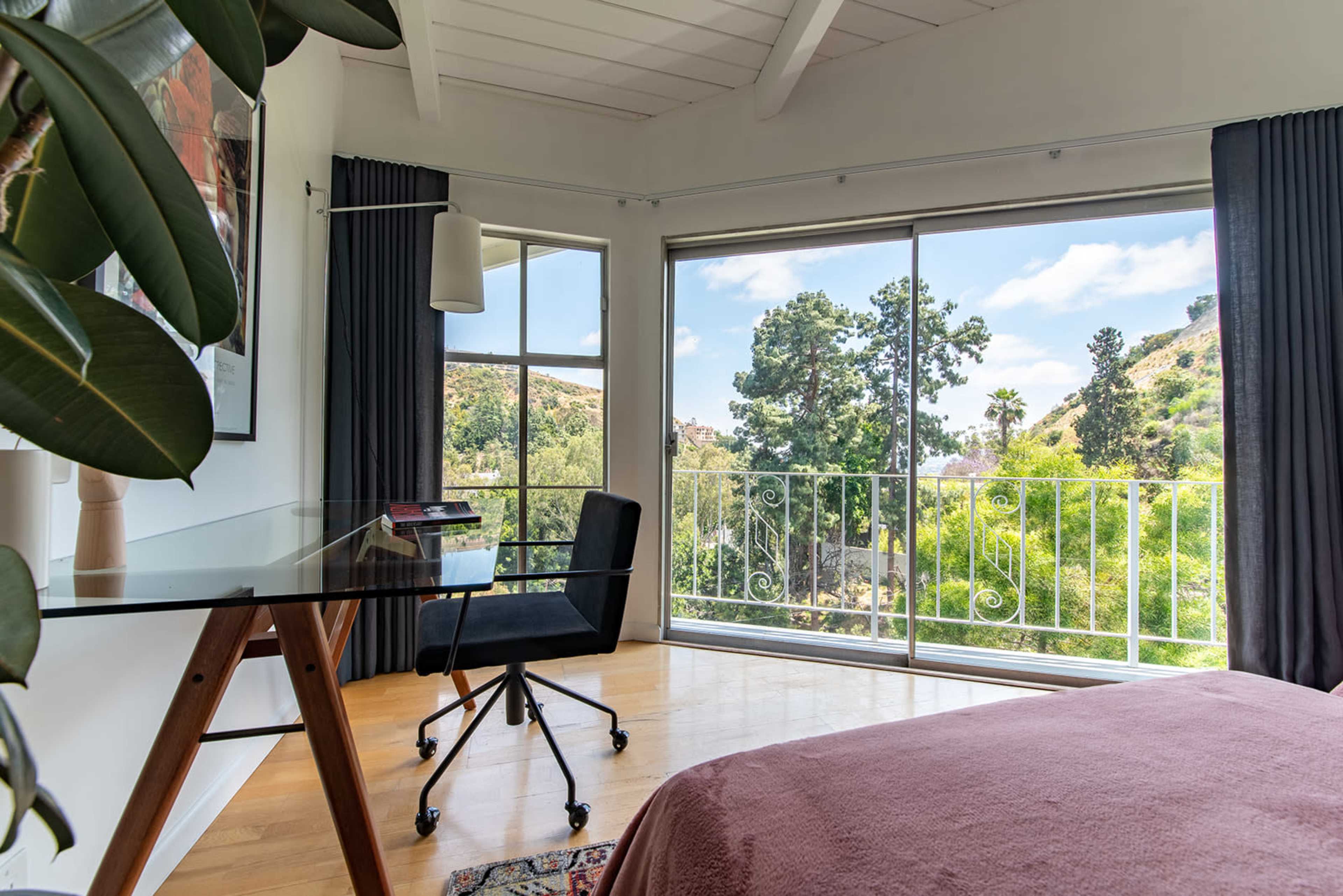 A bright bedroom features a glass balcony door overlooking a green landscape, with a desk and chair positioned nearby.
