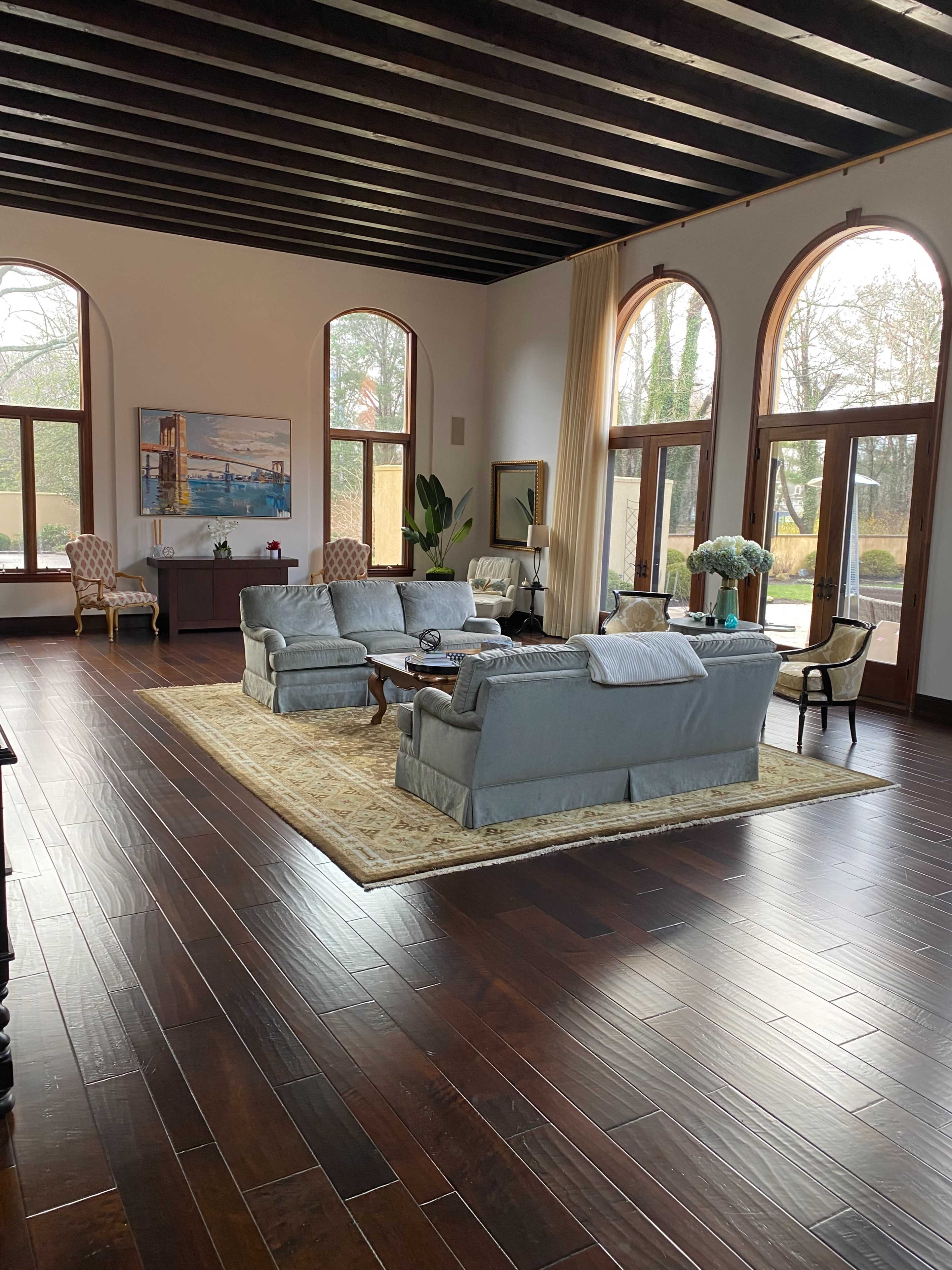 A spacious living room with a light blue sectional sofa, wooden beams on the ceiling, and large arched windows allowing natural light to enter.