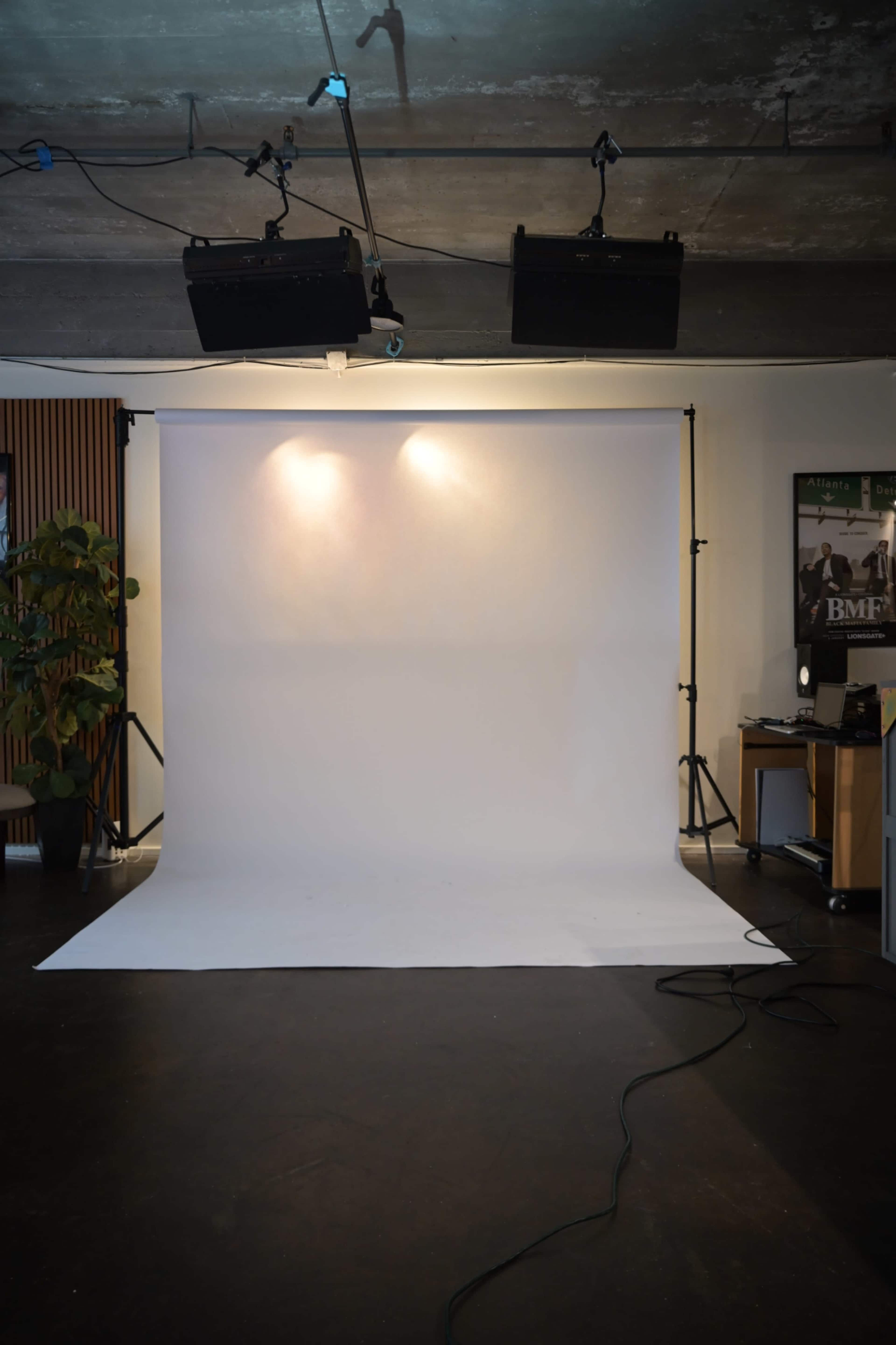 The image shows a photography studio setup with a white backdrop, lighting equipment above, and a small desk on the side.