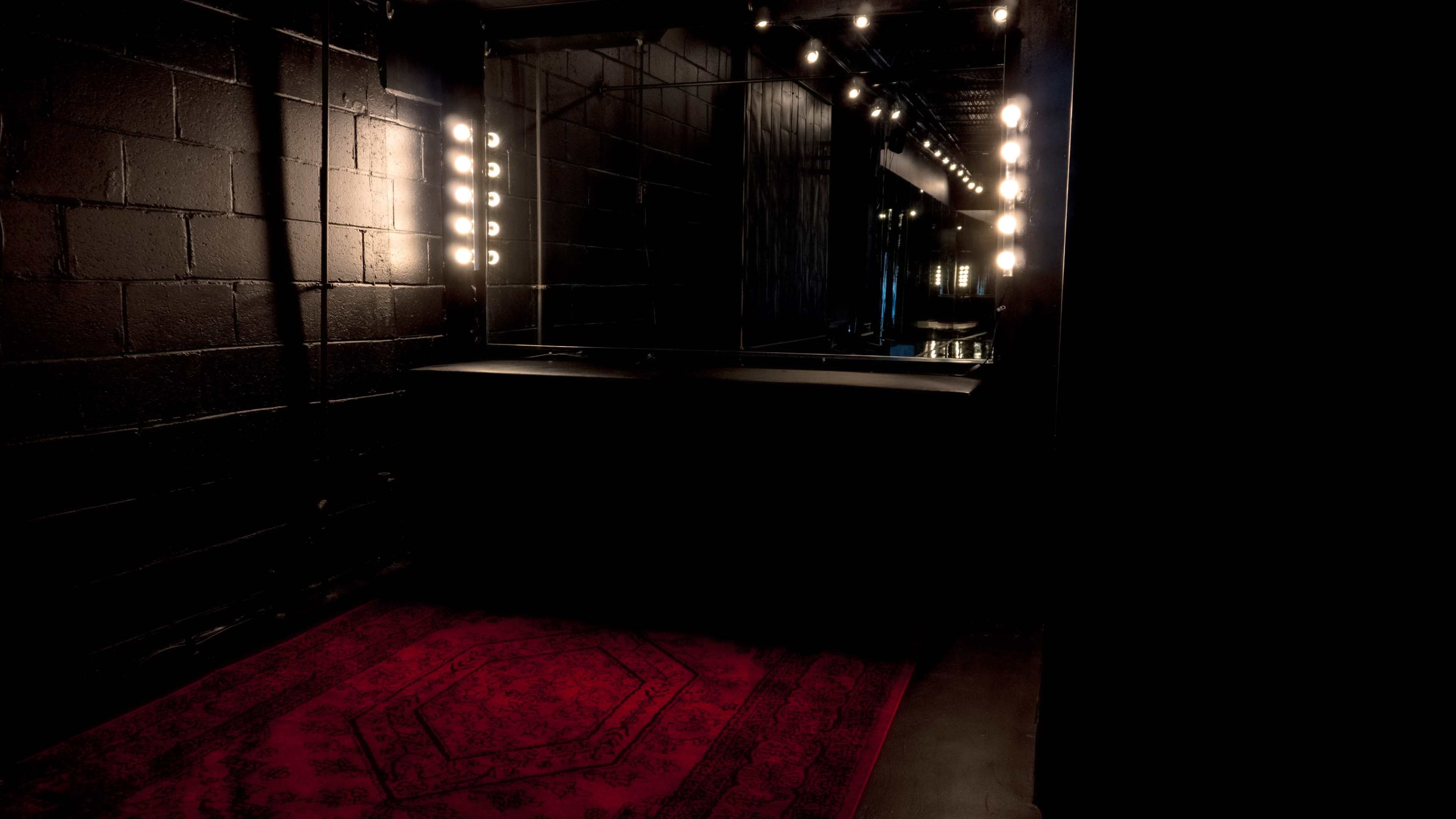 A dimly lit dressing room features a black wall, a large mirror with bright light bulbs, and a red area rug on the floor.