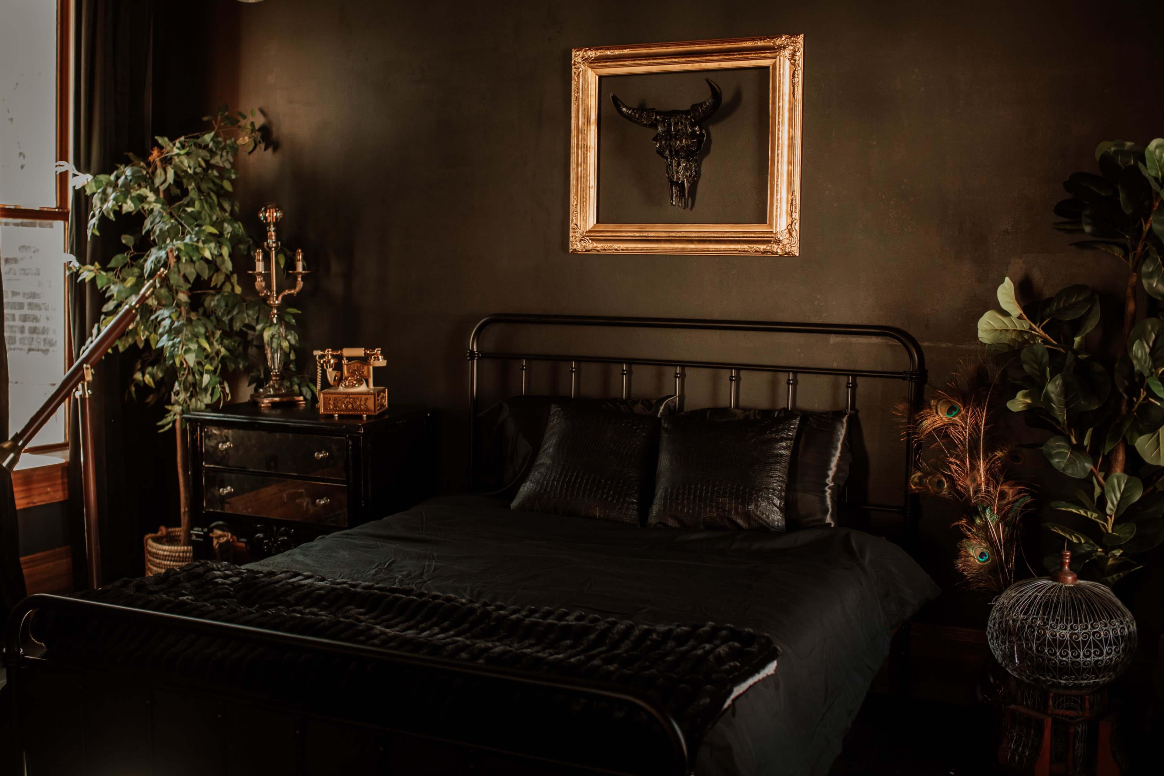 A dark-themed bedroom featuring a metal frame bed with black bedding, a golden-framed bull skull artwork on the wall, and various decorative plants and furnishings.