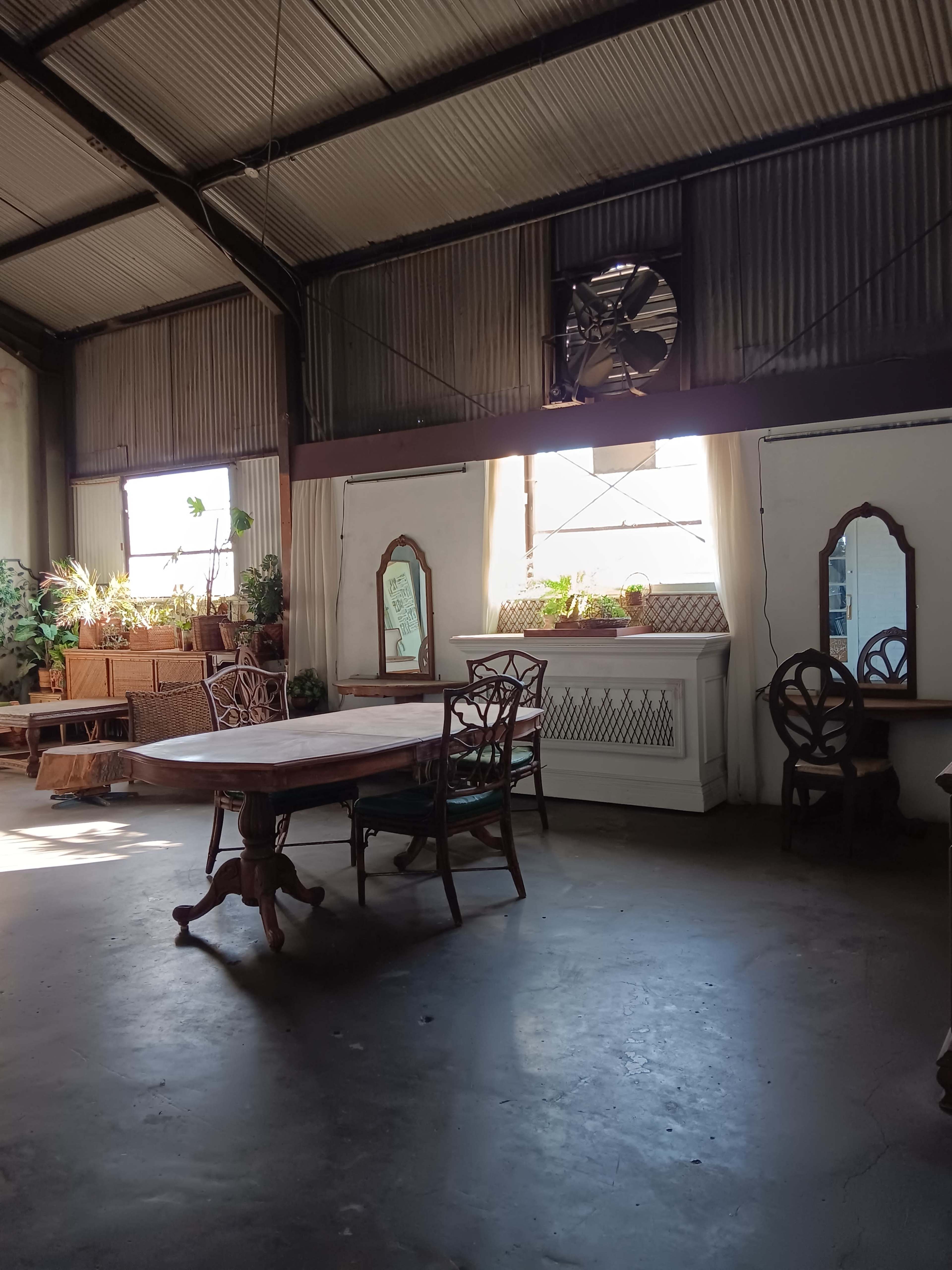 The image shows a spacious industrial-style room with wooden tables, chairs, and large windows allowing natural light to filter in, accompanied by potted plants and decorative mirrors.