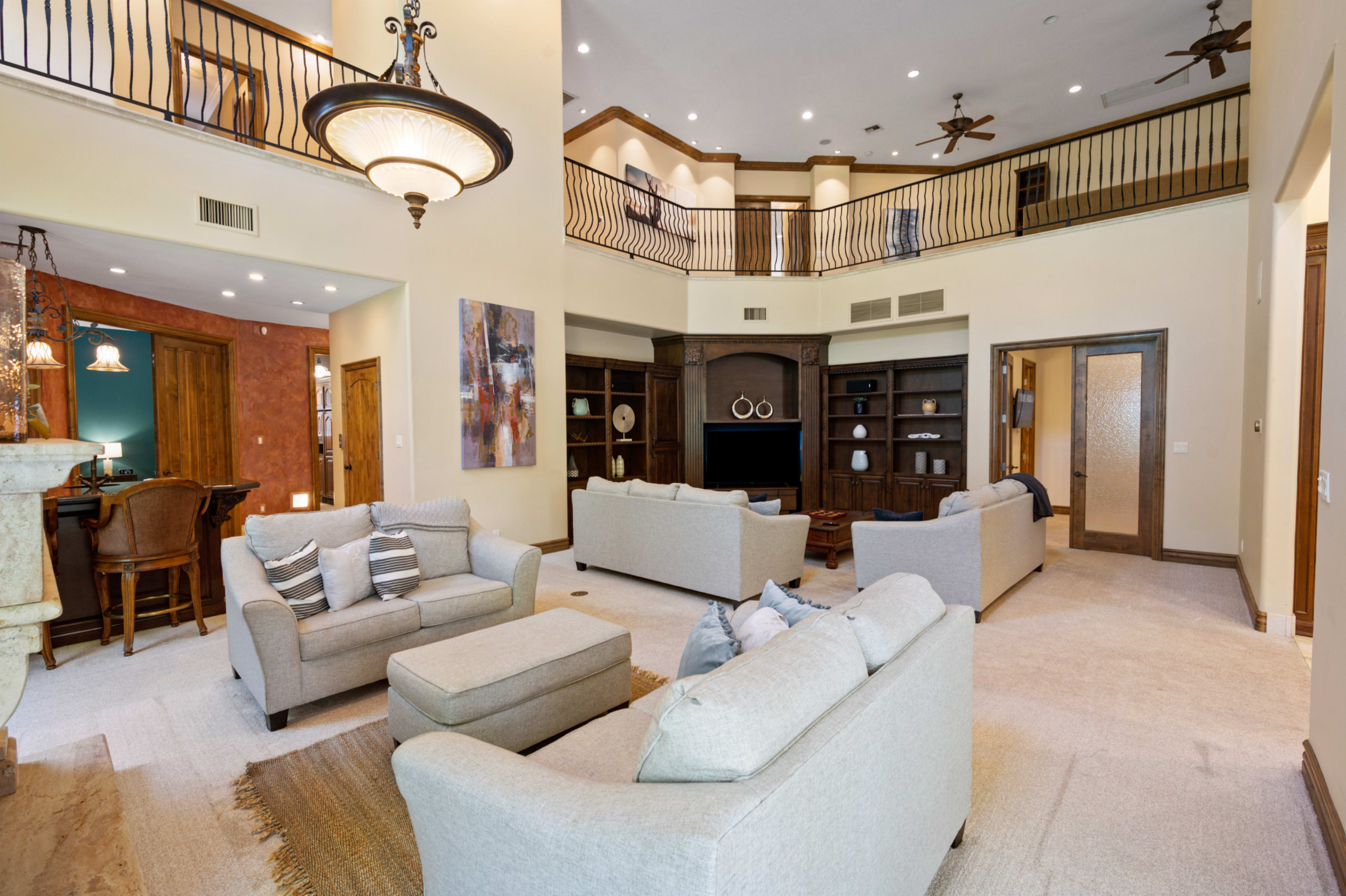 The image shows a spacious living room with two light-colored sofas, a large entertainment unit, and a high ceiling with a balcony above.