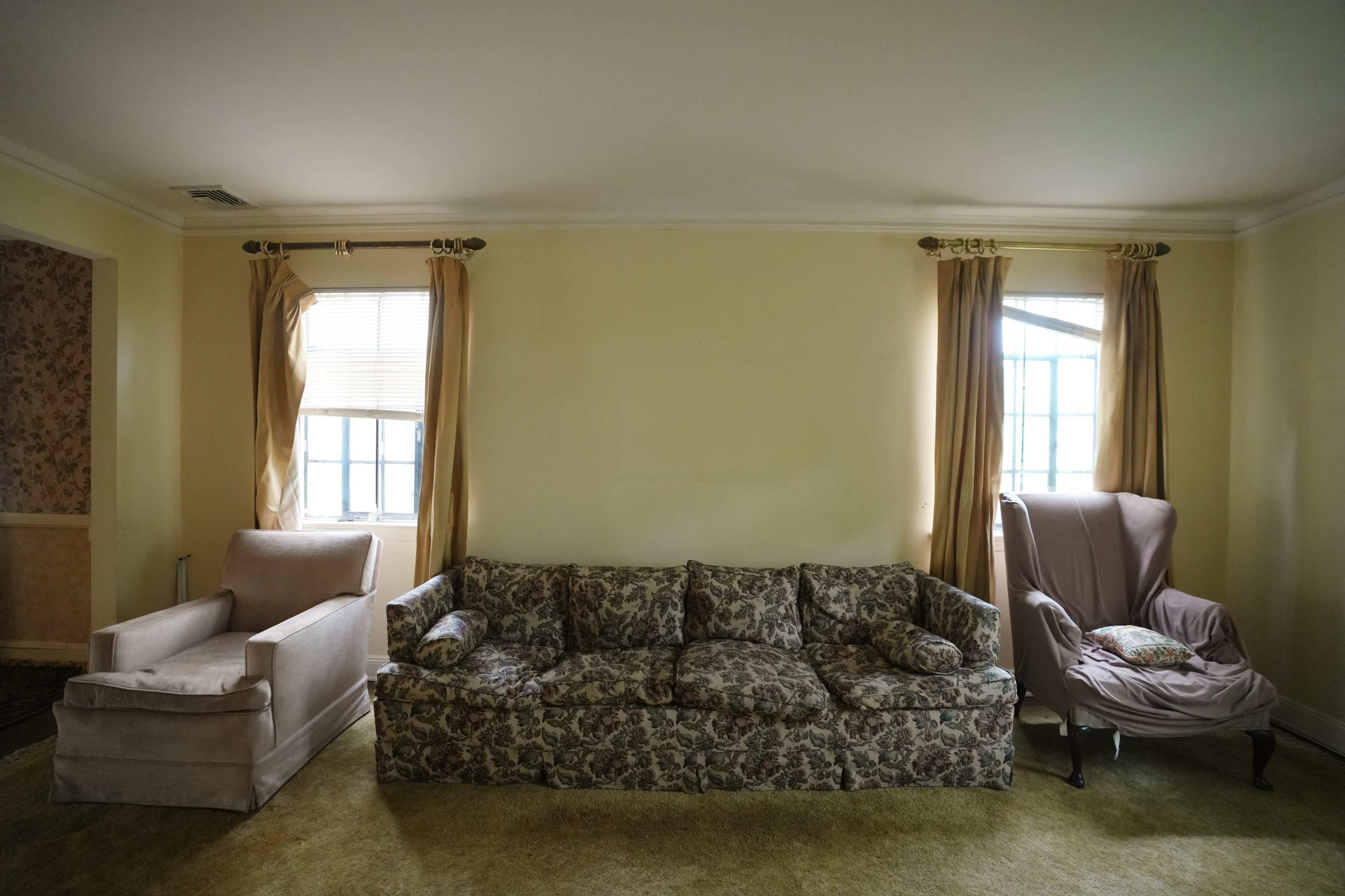 The photo shows a living room with a floral-patterned couch, a chaise lounge, and a wingback chair, all set against yellow walls and windows with light curtains.