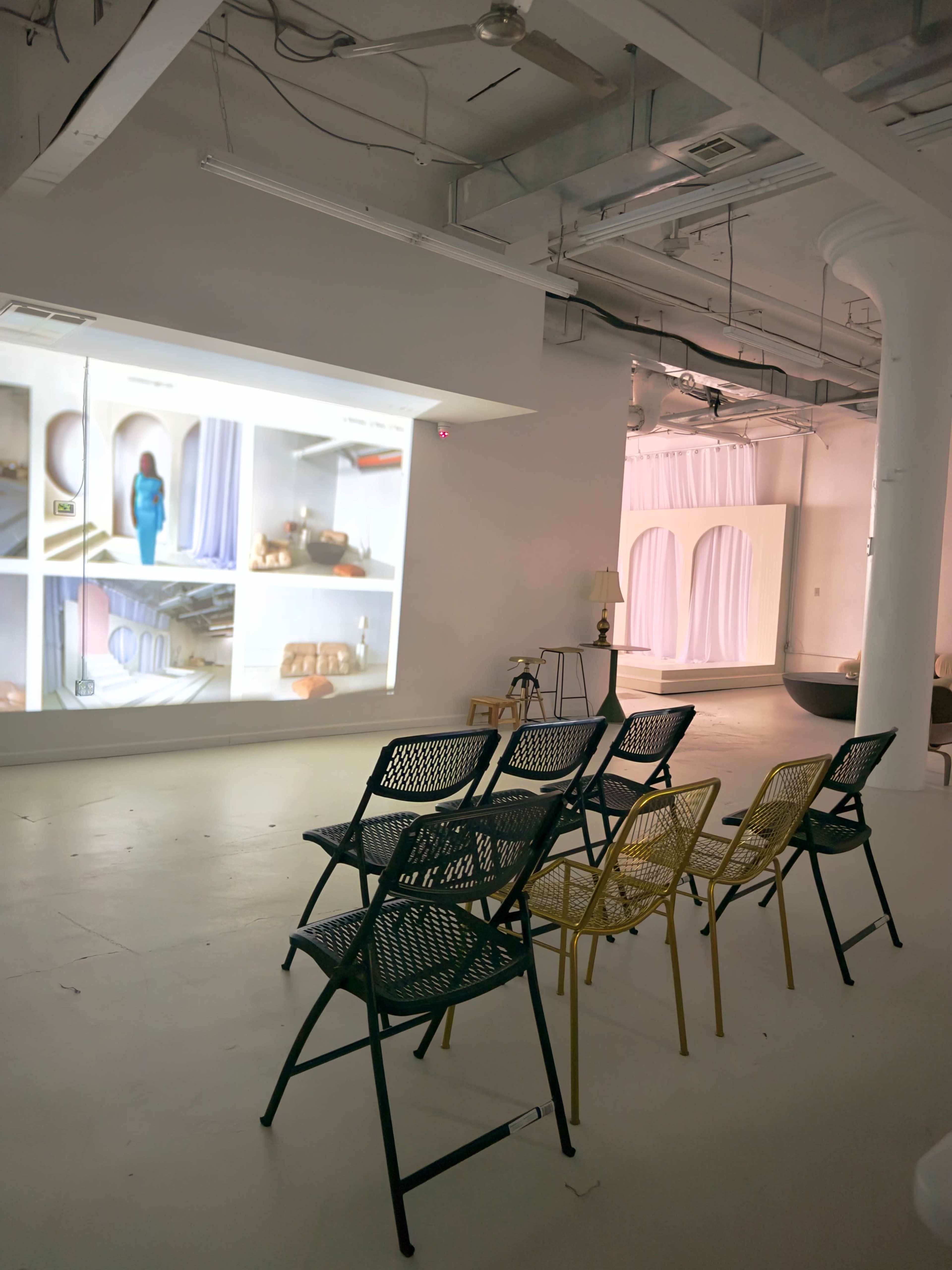 A minimalistic gallery space features a projection displaying images of interiors, with several chairs arranged in front for viewing.