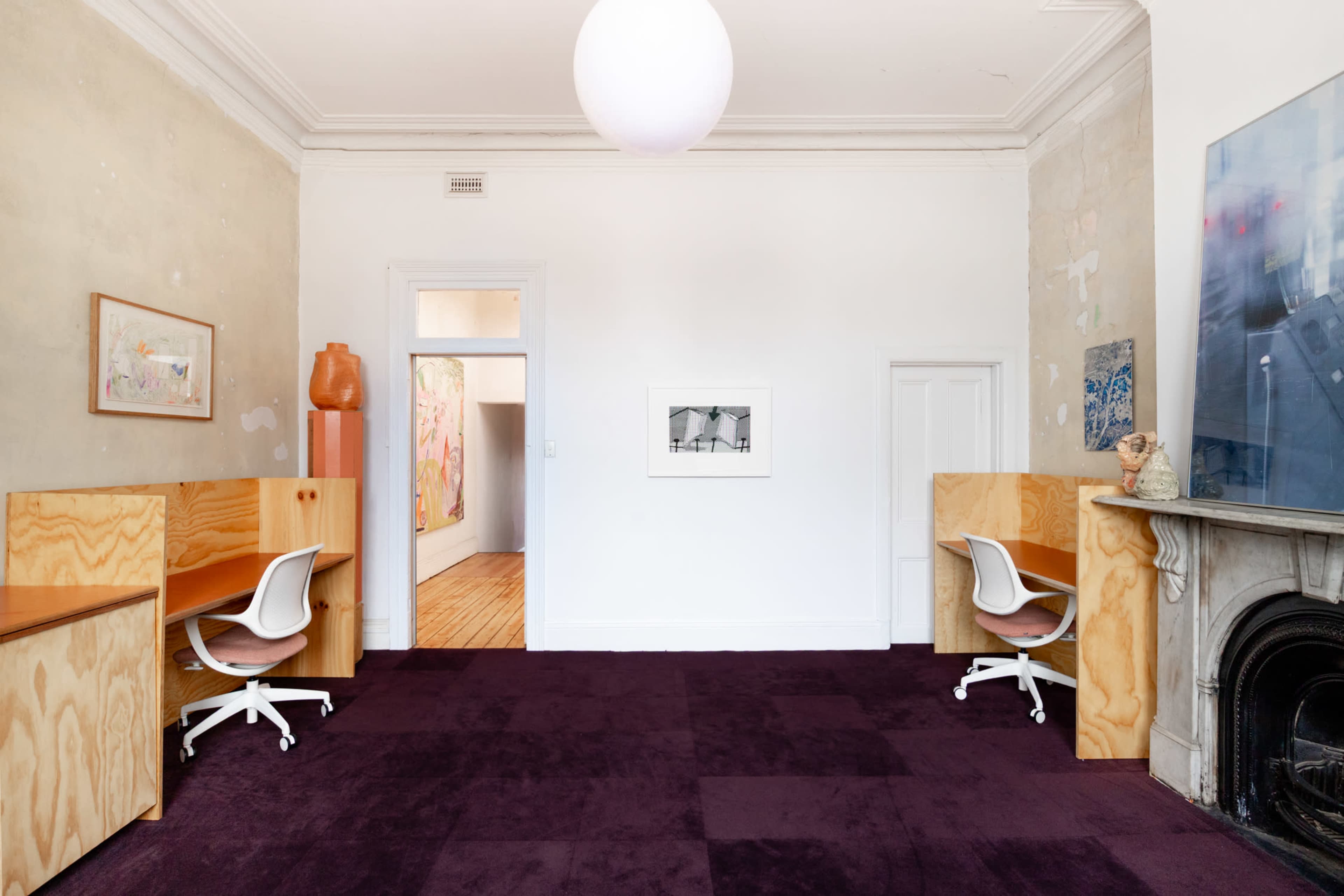 The image depicts a minimally furnished room with two wooden desks and chairs, a large circular light fixture, and a fireplace against the wall, featuring an empty doorway leading to another space.