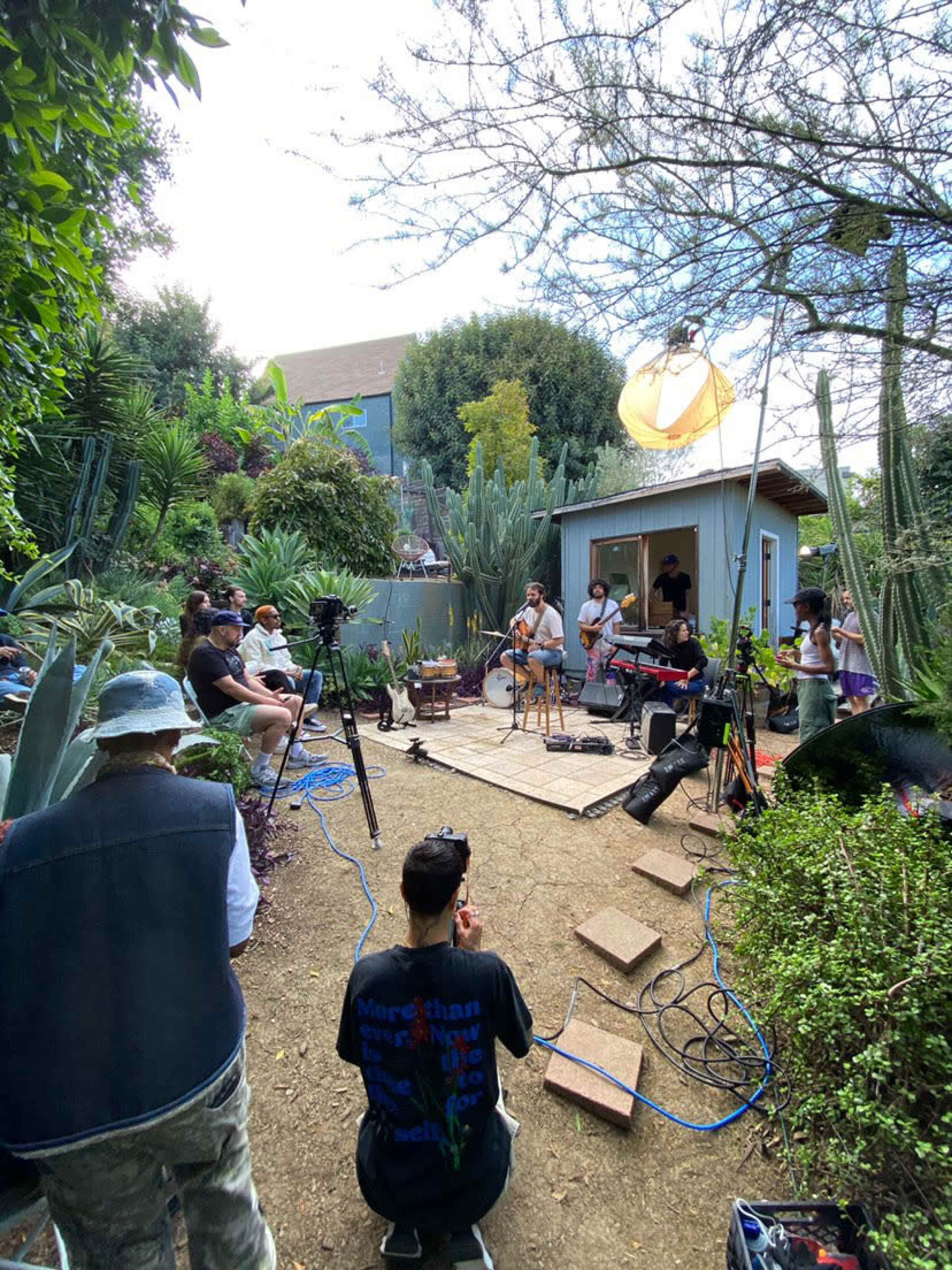 A small outdoor music performance takes place in a garden surrounded by various plants, with an audience seated on the ground and musicians on a wooden stage.