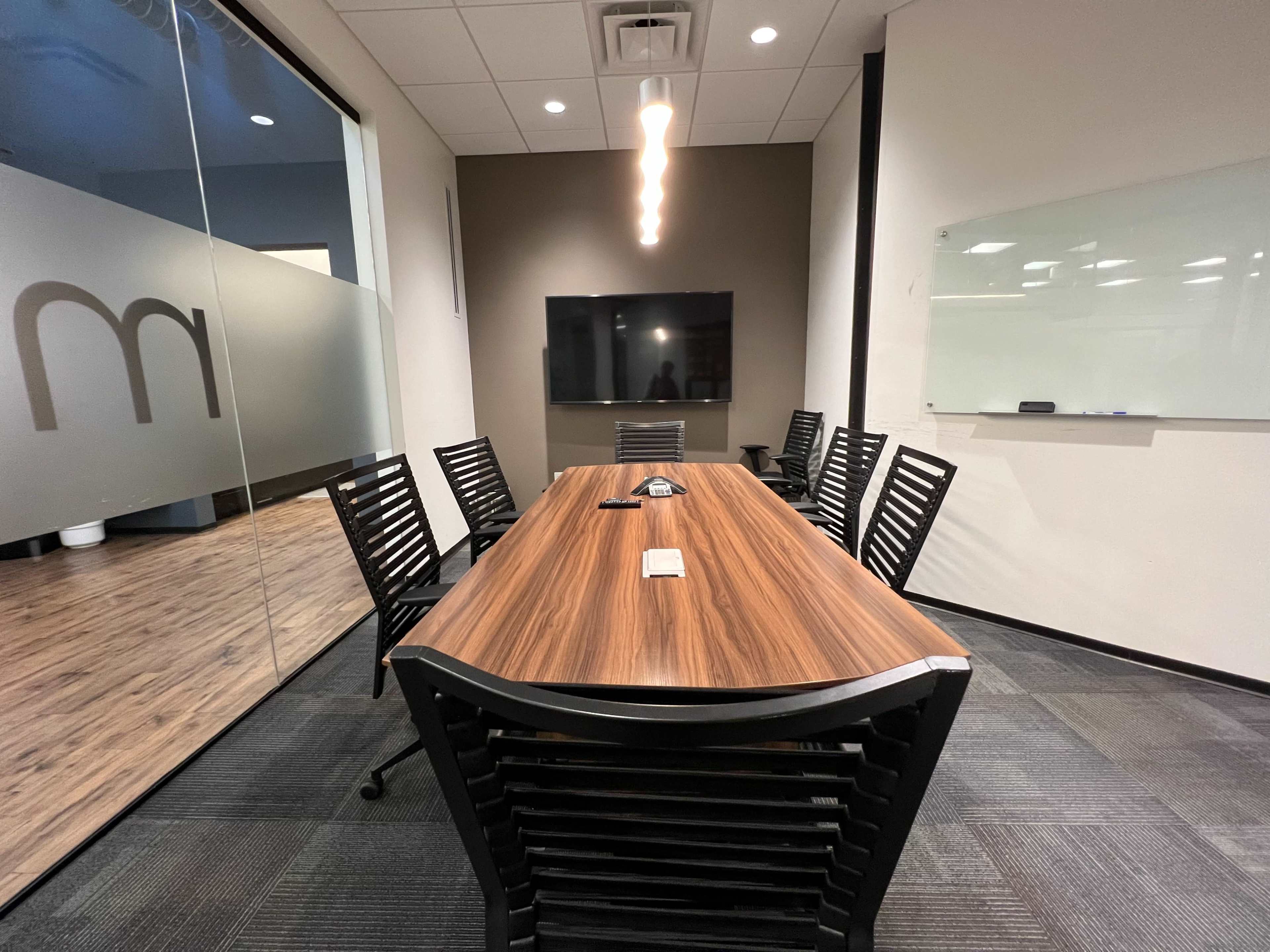 A conference room features a long wooden table surrounded by black chairs, with a large screen and whiteboard on opposite walls.