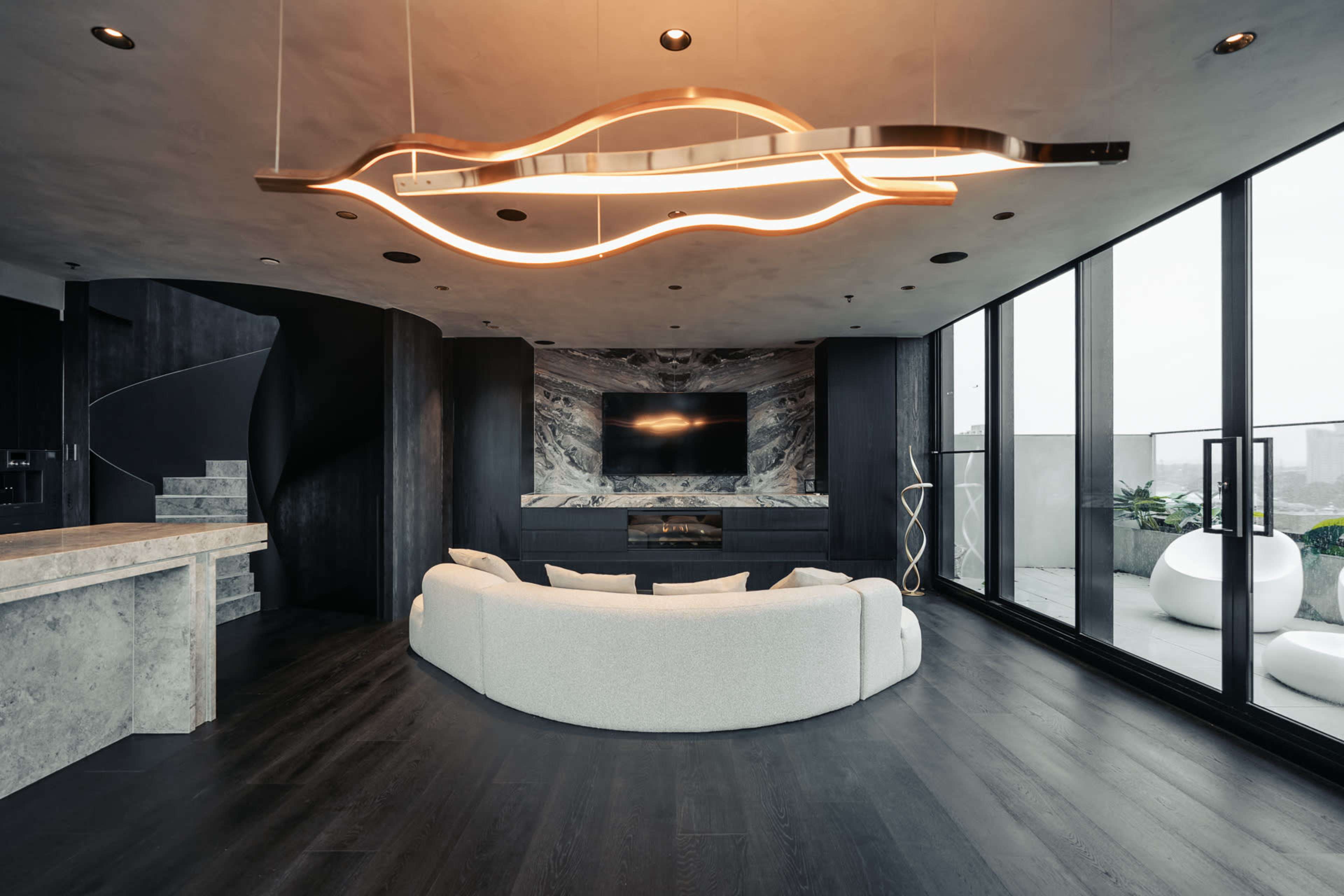 The image shows a modern living room with a curved white sofa, a dark accent wall featuring a television, and large windows that overlook an outdoor space.