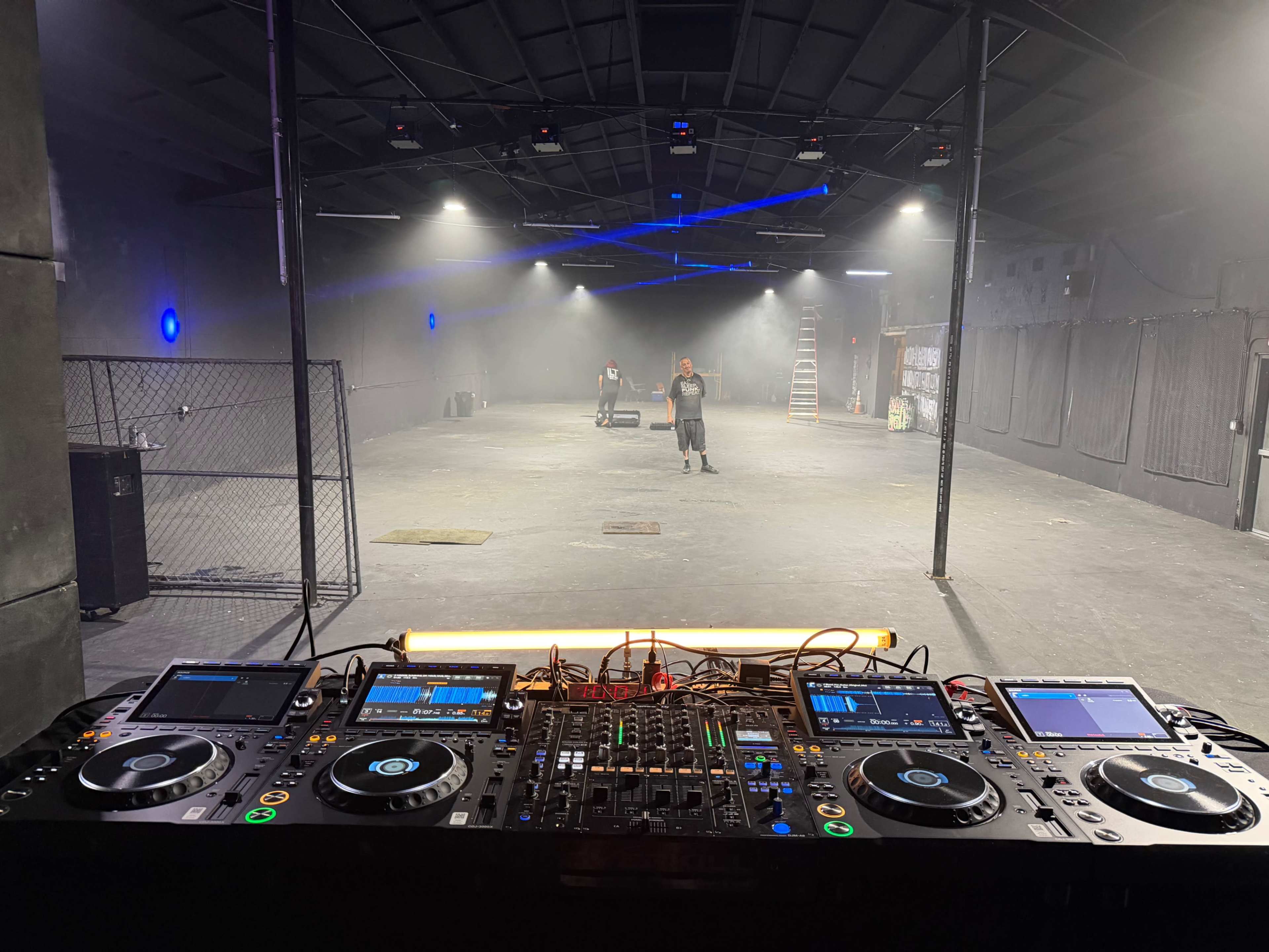 The image shows a DJ setup with controllers and mixers in the foreground, facing an empty warehouse space illuminated by soft lighting and smoke effects.