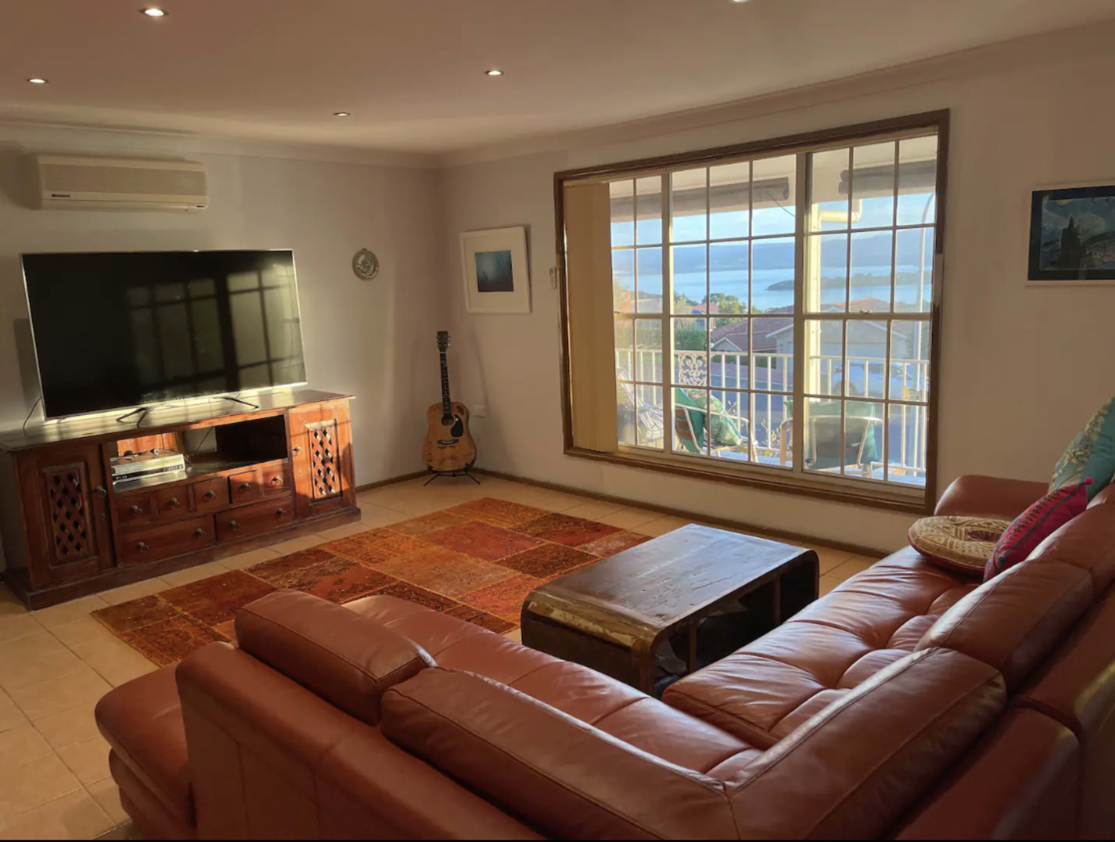 The living room features a large television on a wooden cabinet, a comfortable leather sofa, an acoustic guitar by the window, and a coffee table on a patterned rug.