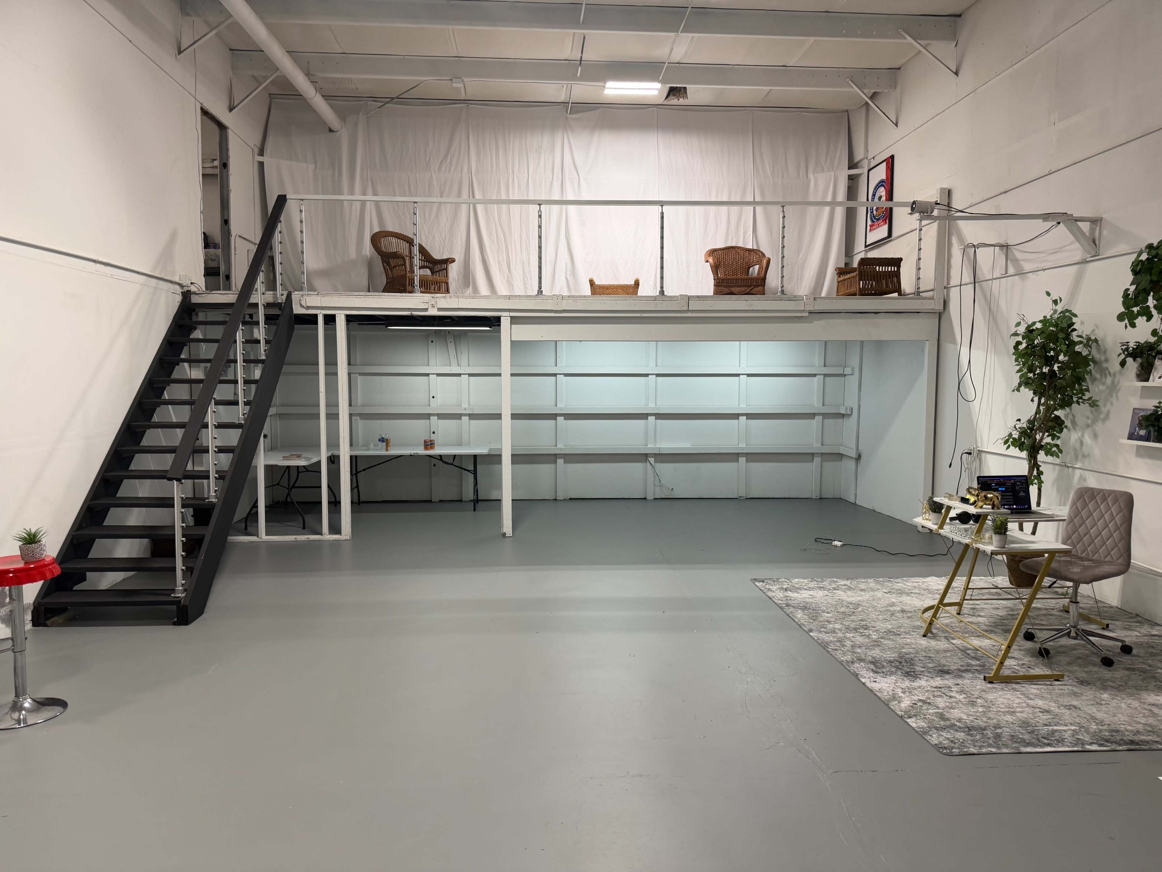 The image shows an industrial-style room with a loft area, featuring a staircase, two wicker chairs on the upper level, and a workspace setup with a desk and a chair on the ground floor.
