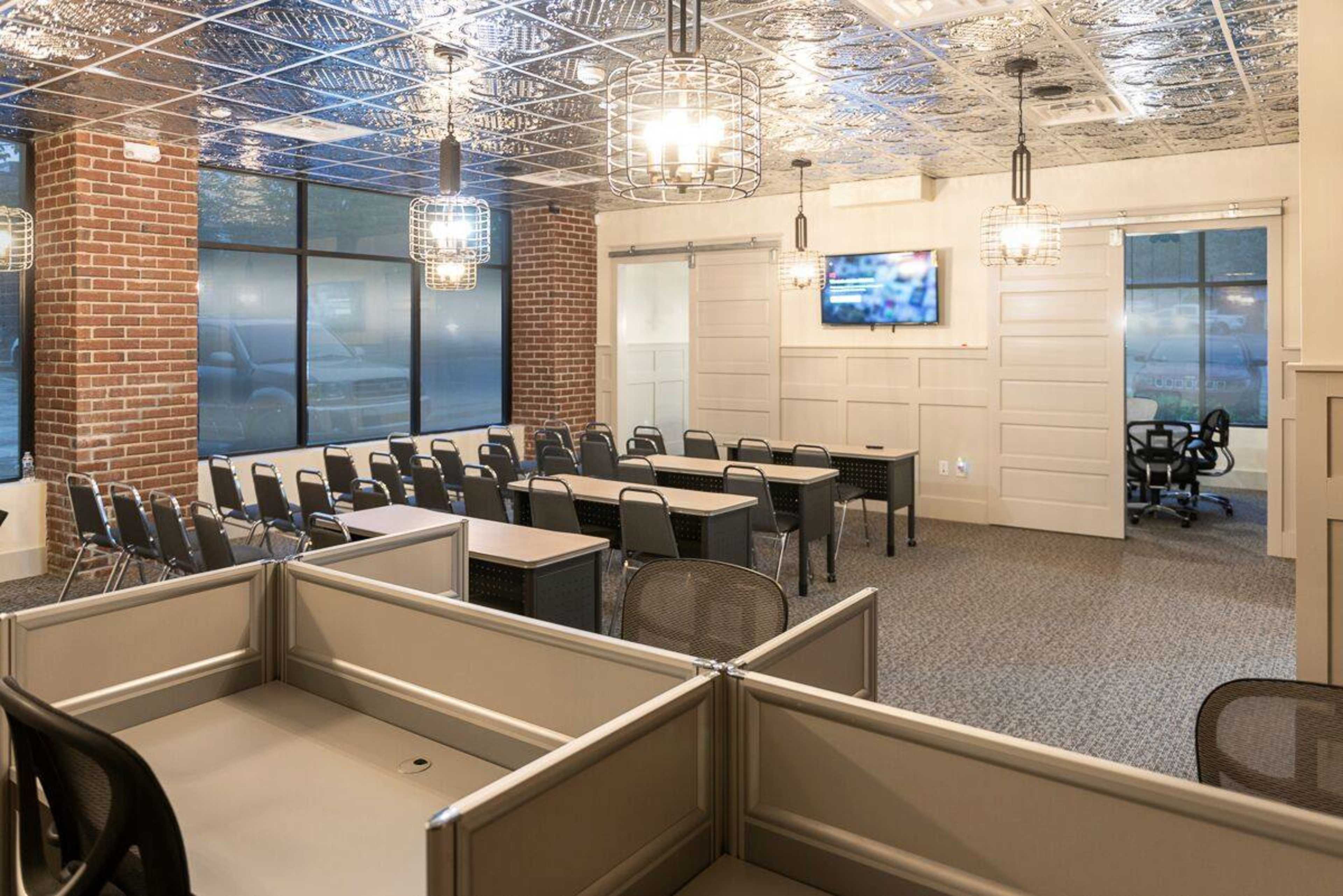 A modern office space with a mix of individual workstations and a conference area featuring rows of tables and chairs, along with a television mounted on the wall.