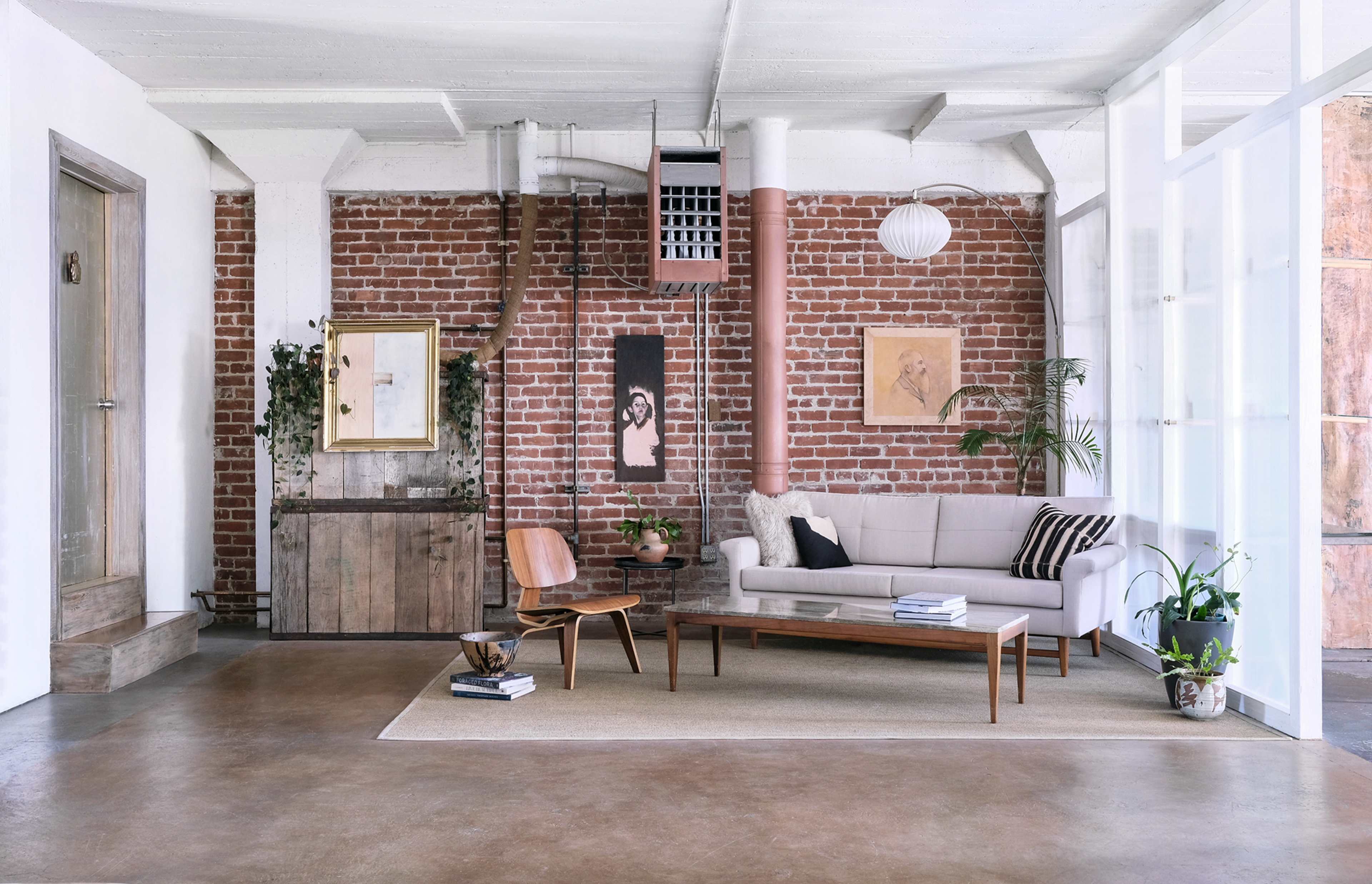 A modern living room with exposed brick walls, a light beige sofa, a wooden coffee table, and a mix of plants and artwork decorating the space.
