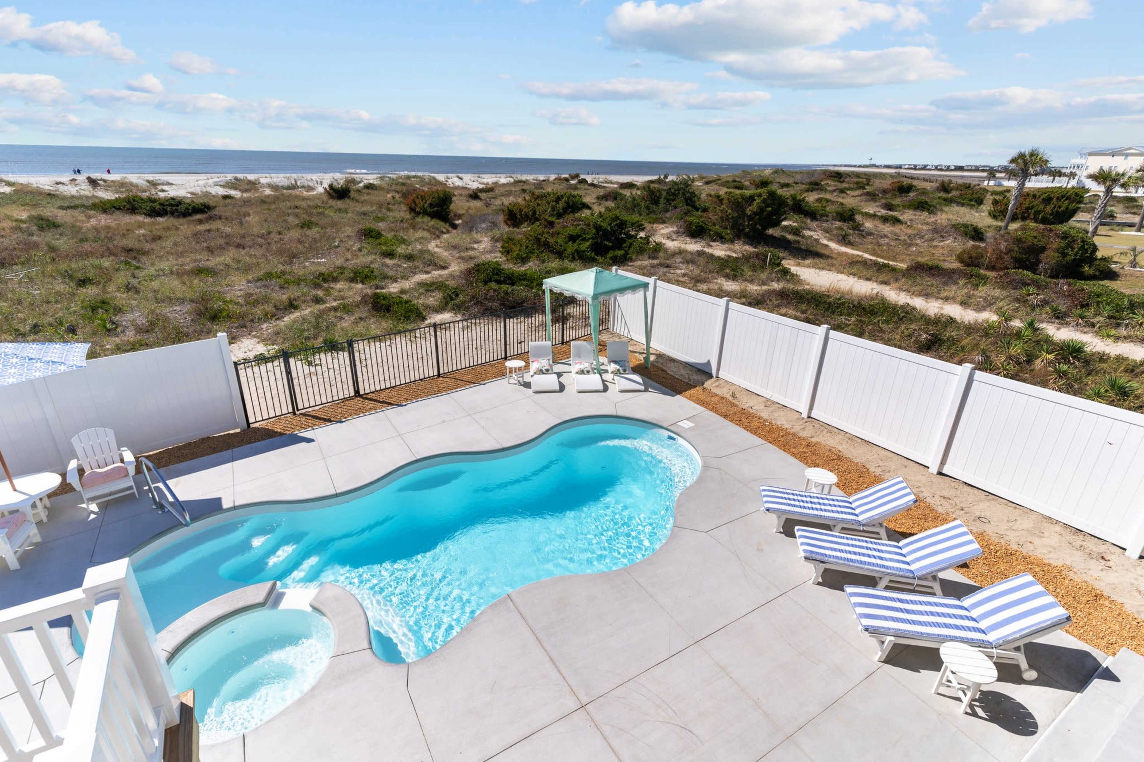 A fenced pool area with lounge chairs, a hot tub, and a view of the beach and greenery in the background.