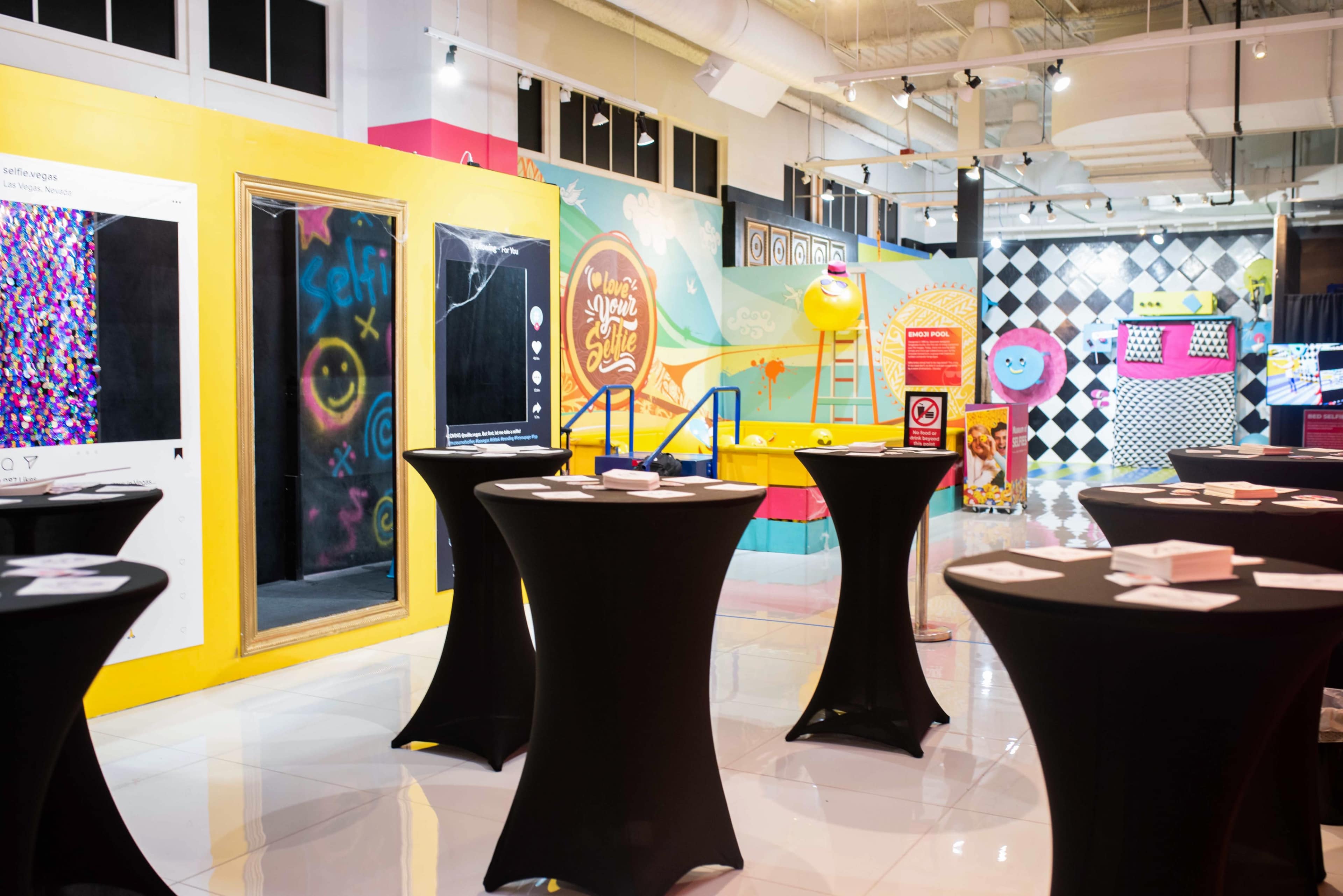 A brightly colored exhibition space featuring black tables, vibrant wall art, and interactive installations.