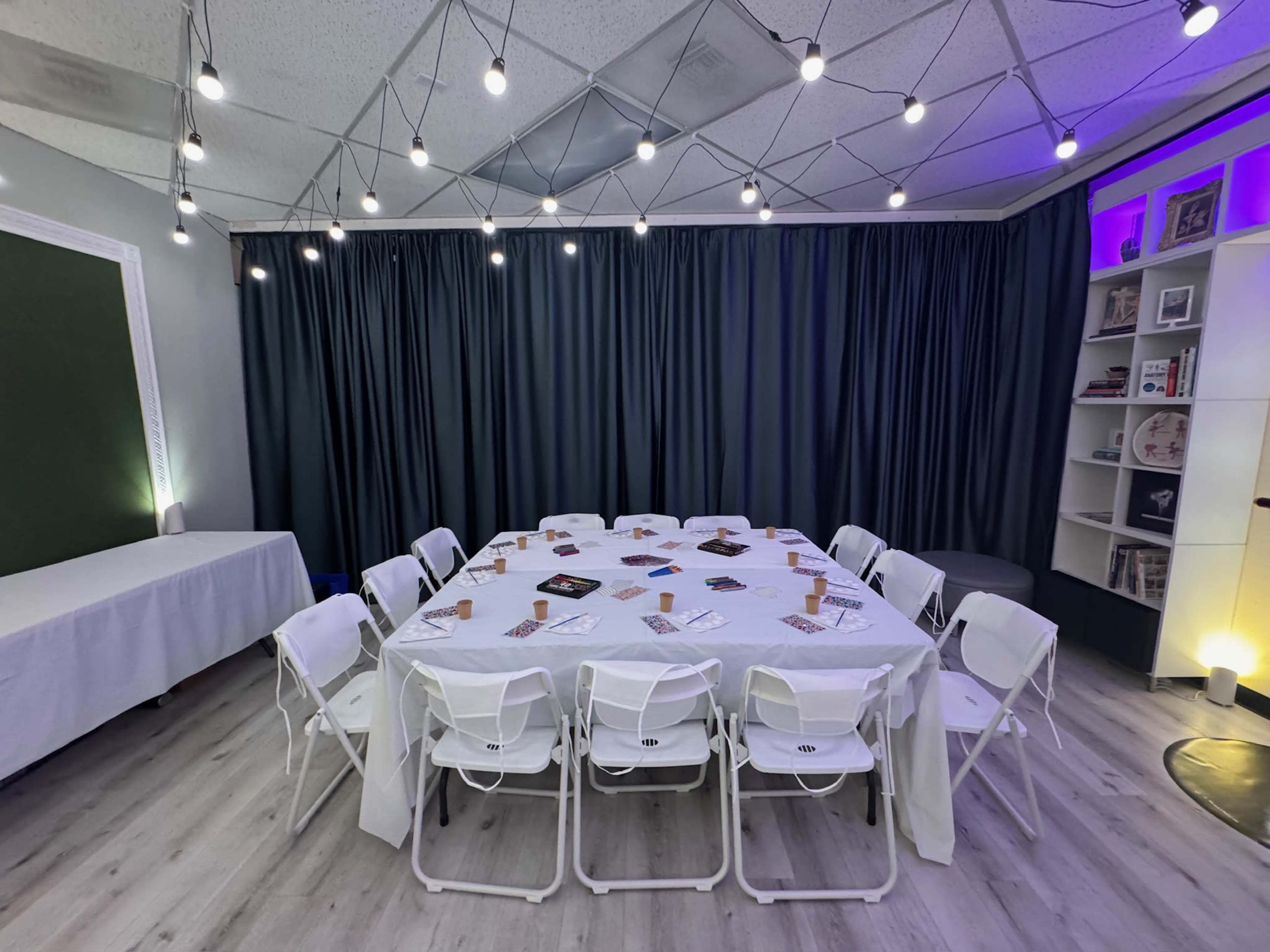 A round table covered with a white tablecloth is surrounded by white folding chairs in a well-lit room with dark curtains and overhead lights.