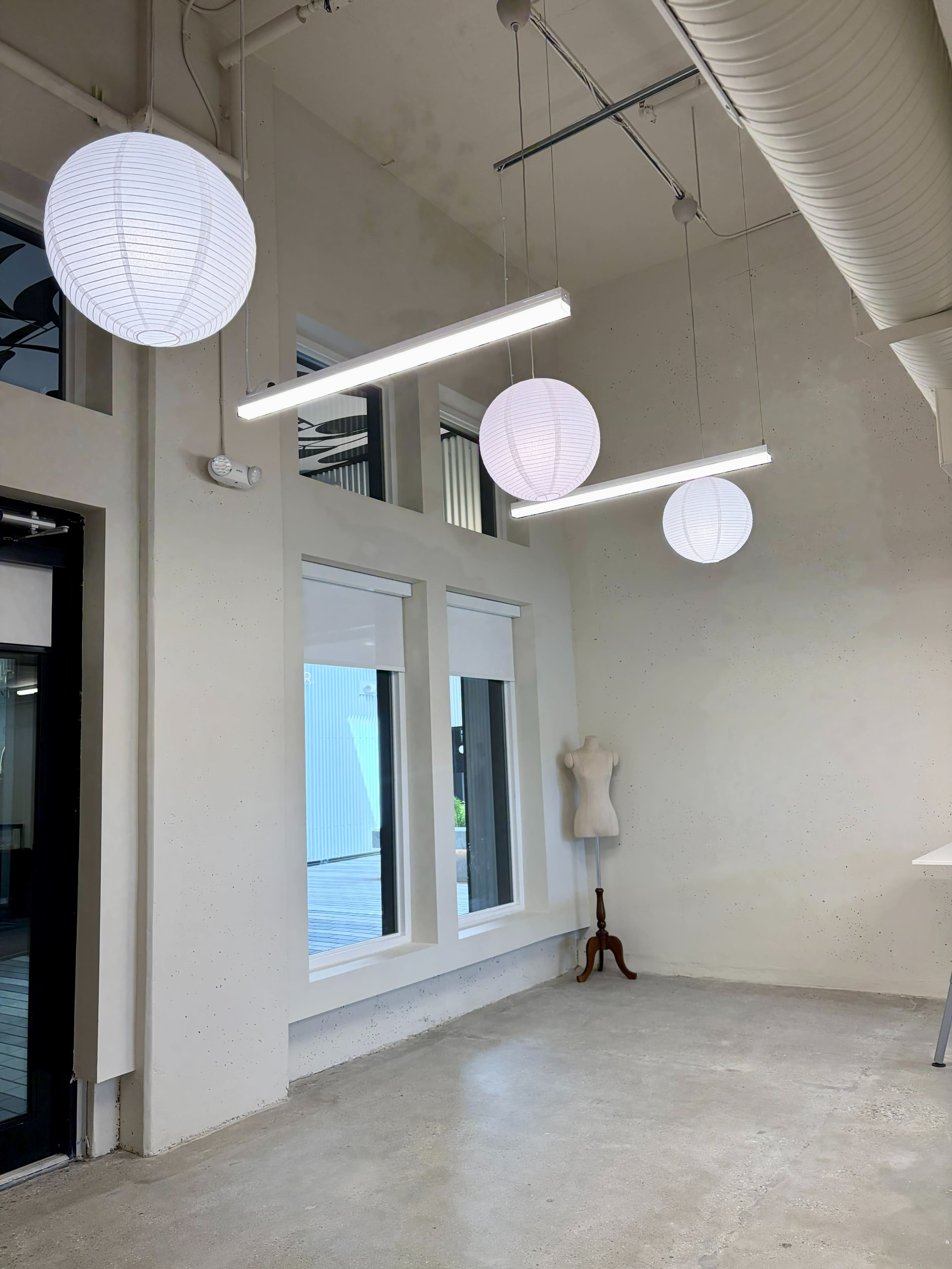 The image shows a minimalist interior with three hanging light fixtures and a mannequin against a light-colored wall, featuring large windows that let in natural light.