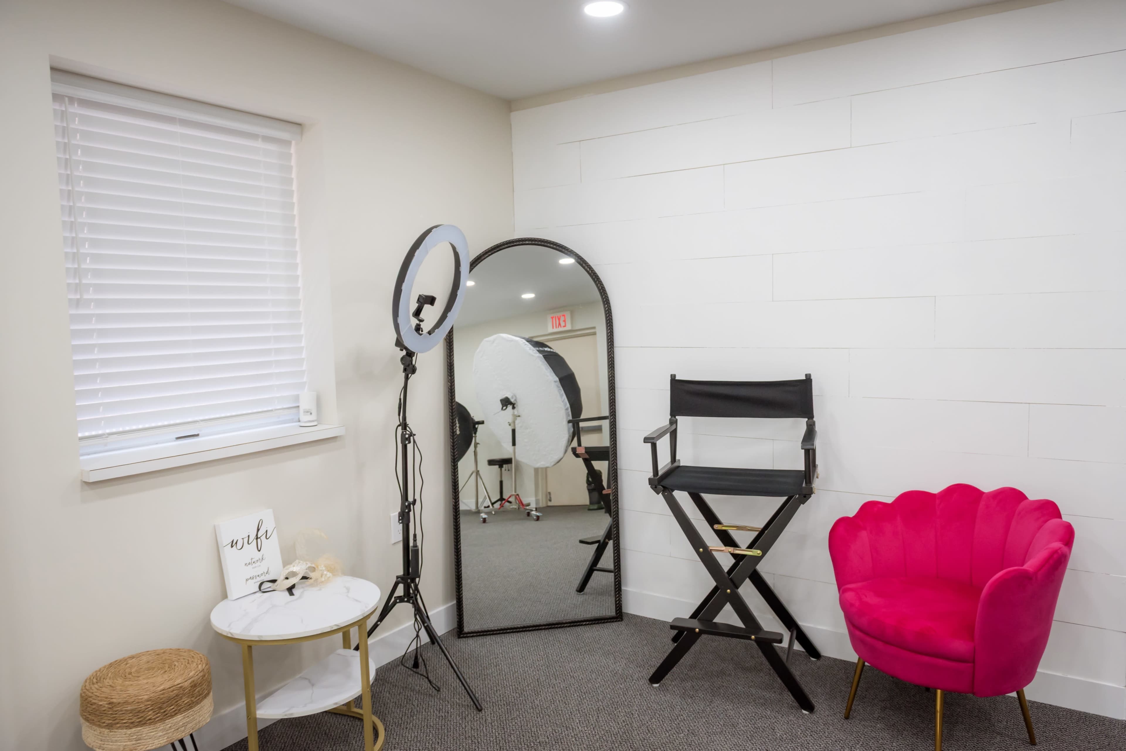 The image shows a well-lit room featuring a large mirror, a black director's chair, a pink accent chair, and photography lighting equipment.