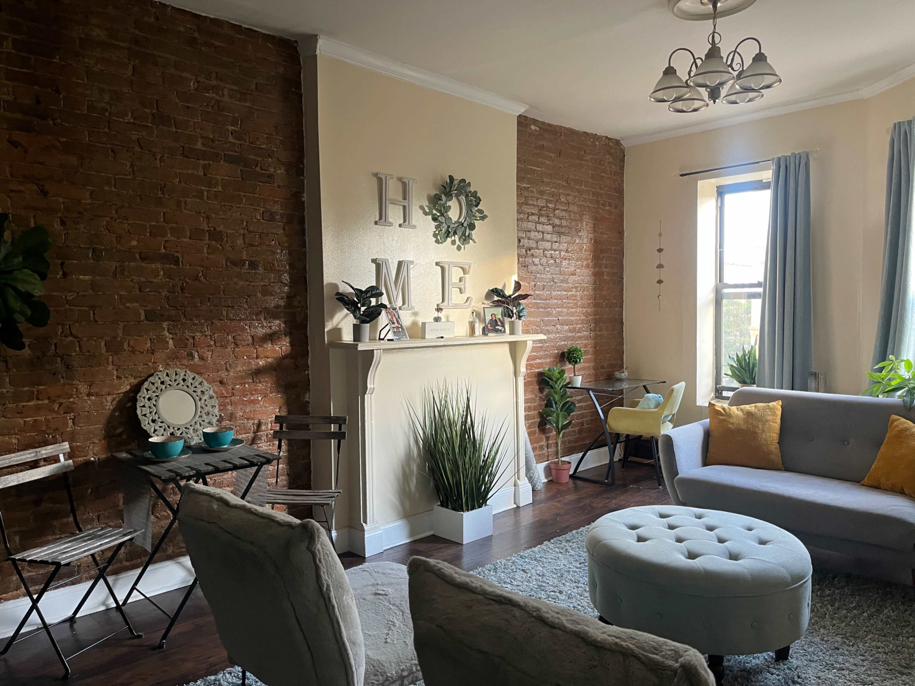 A cozy living room with exposed brick walls, a decorative fireplace, a circular ottoman, and potted plants.