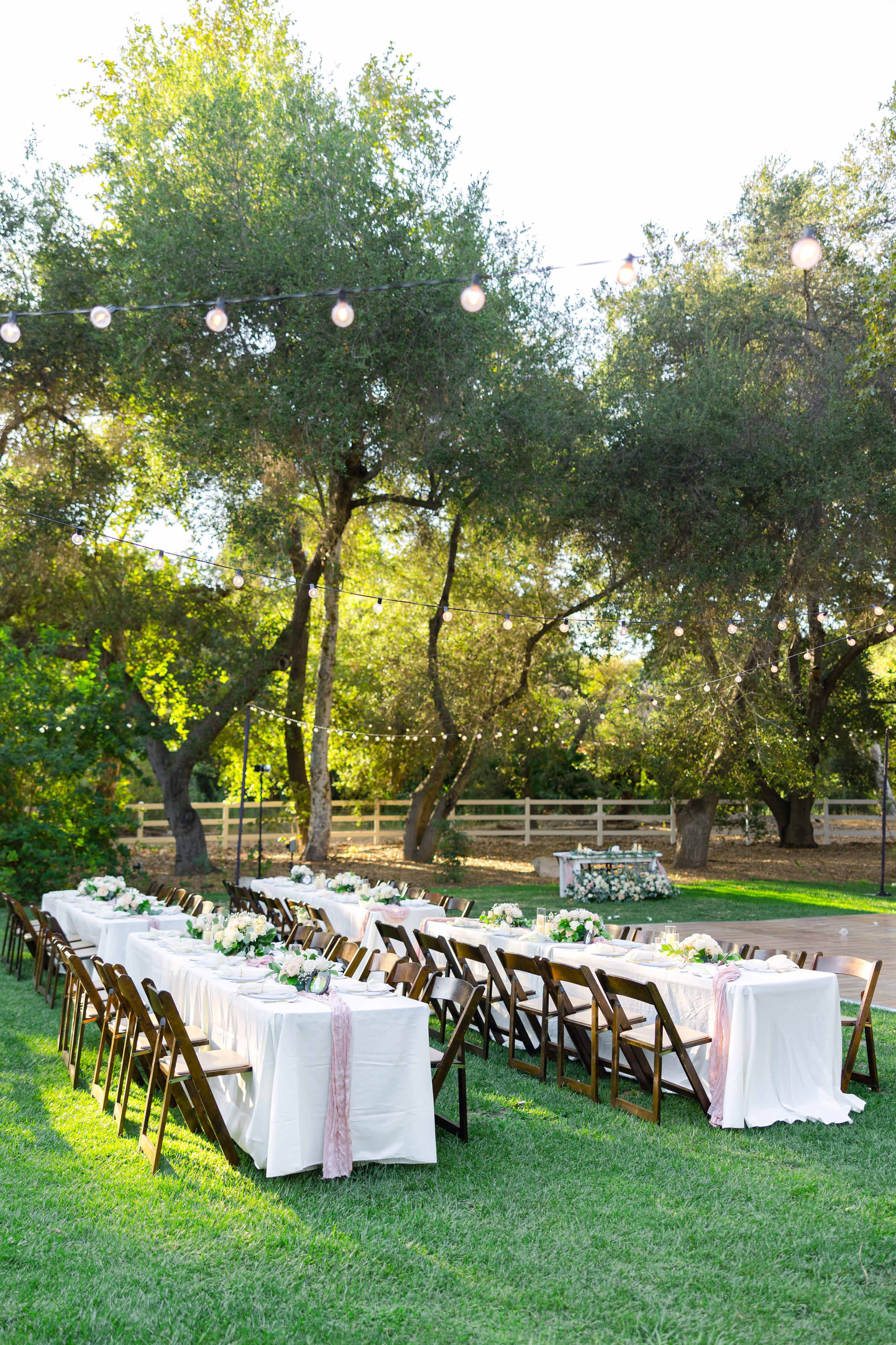 A decorated outdoor dining area features long tables with white tablecloths and floral arrangements under string lights amidst greenery.