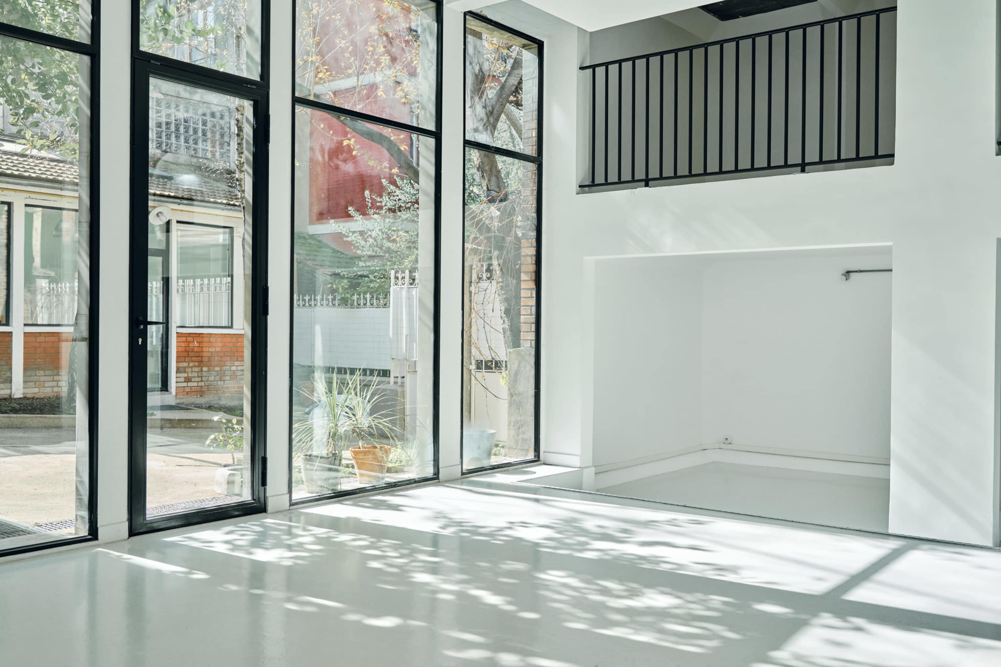 The image shows a bright, open space with large glass windows allowing natural light to fill the room, which features a minimalist design and a view of greenery outside.