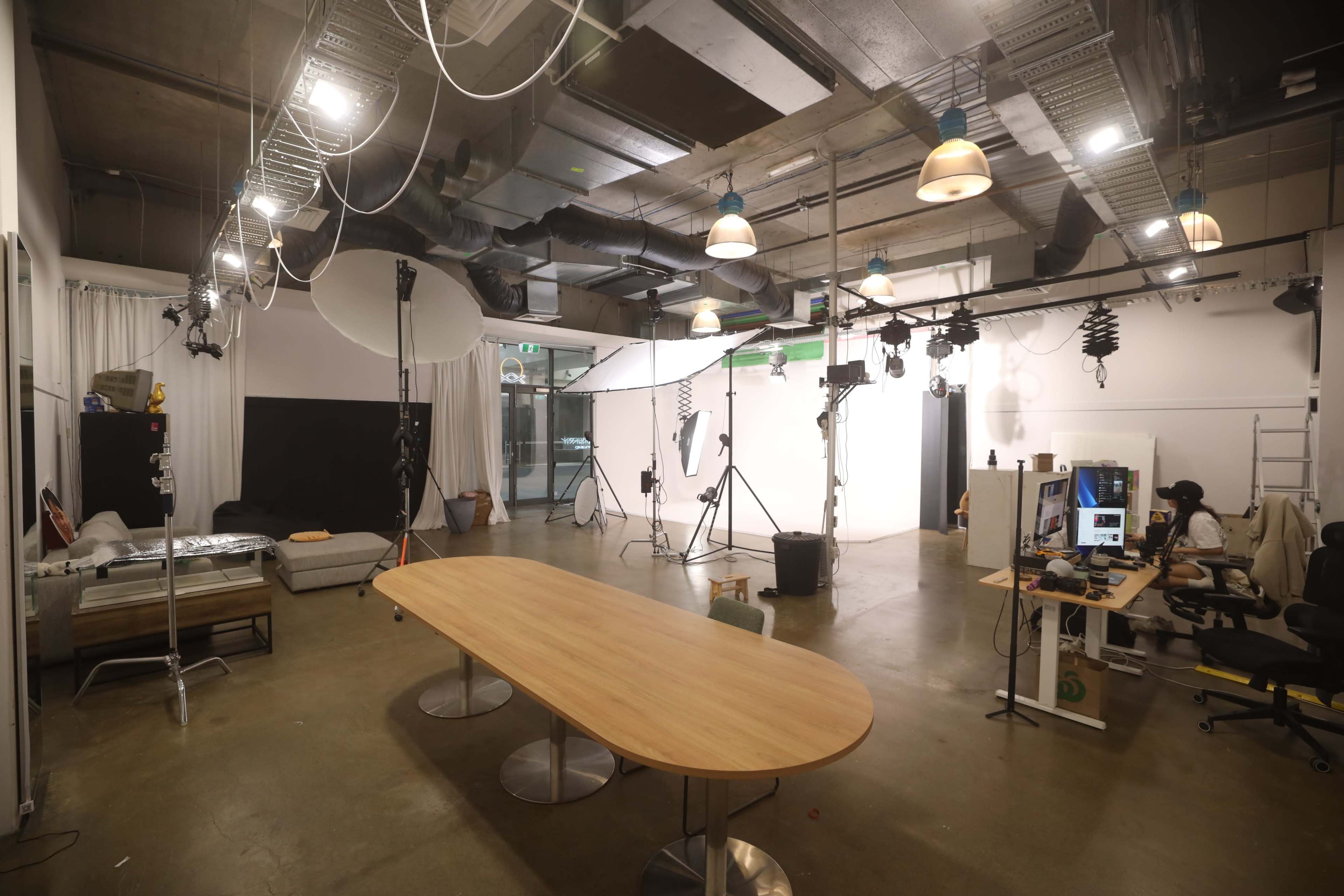 The image shows a spacious studio setup with a large wooden table in the center, multiple lighting fixtures overhead, and various filming equipment positioned around the room.