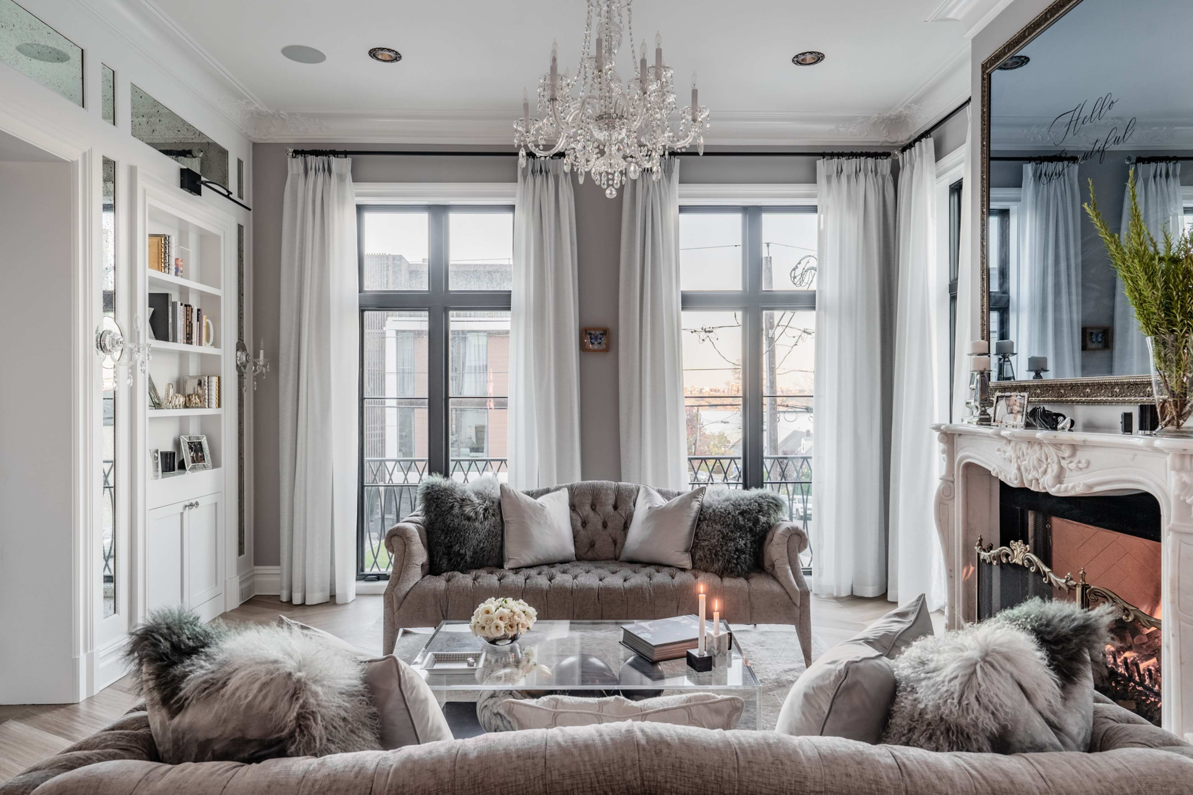 A stylish living room with a chandelier, large windows, a plush sofa, and a decorative fireplace.