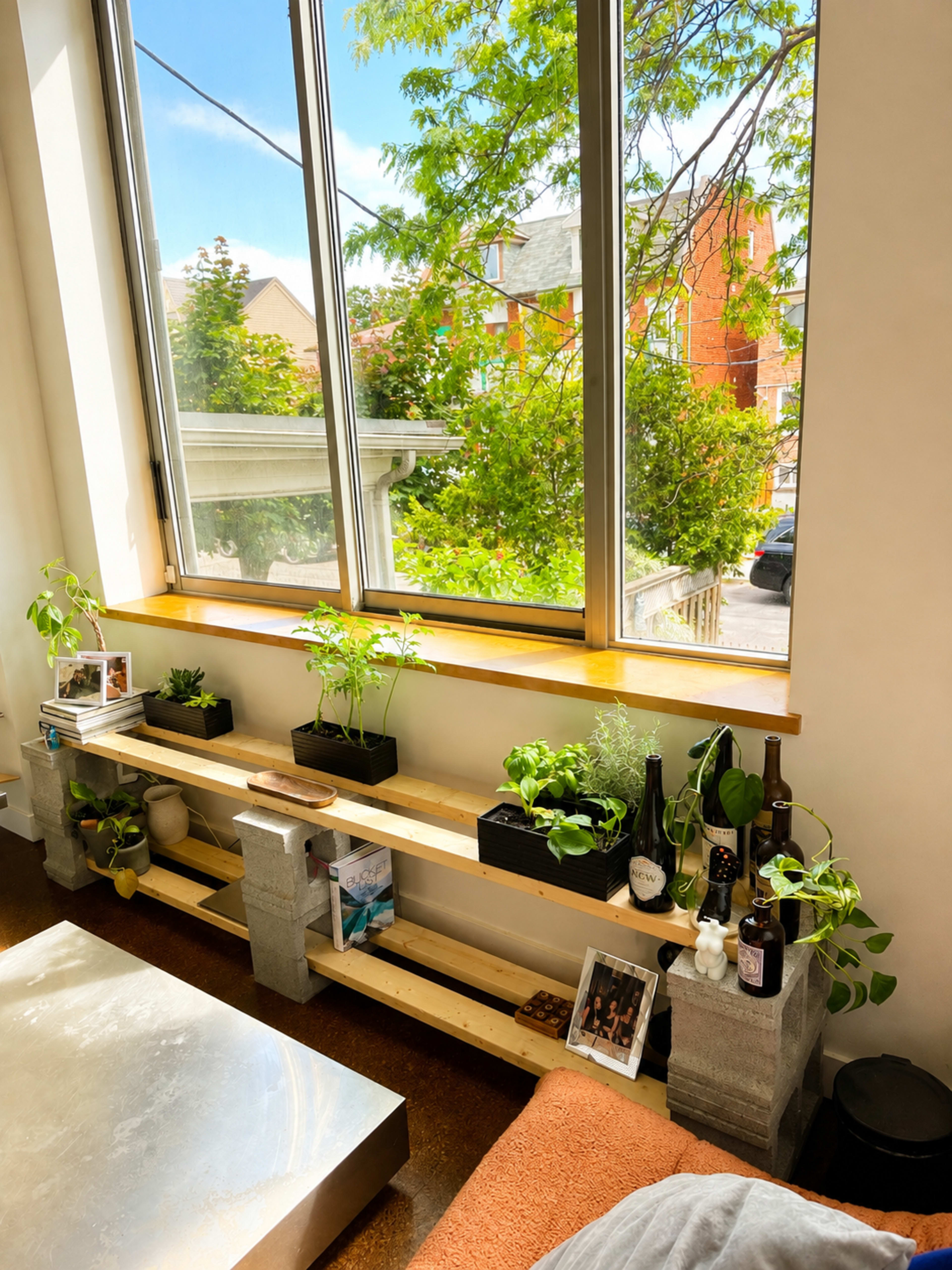 A wooden shelf supported by concrete blocks holds potted plants and decorative items beside large windows that let in natural light.